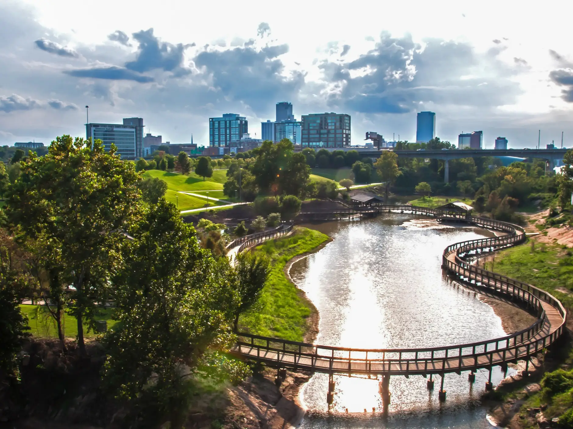 Curving wooden boardwalks cross a reflective river beside green parkland and a city skyline under cloudy skies.