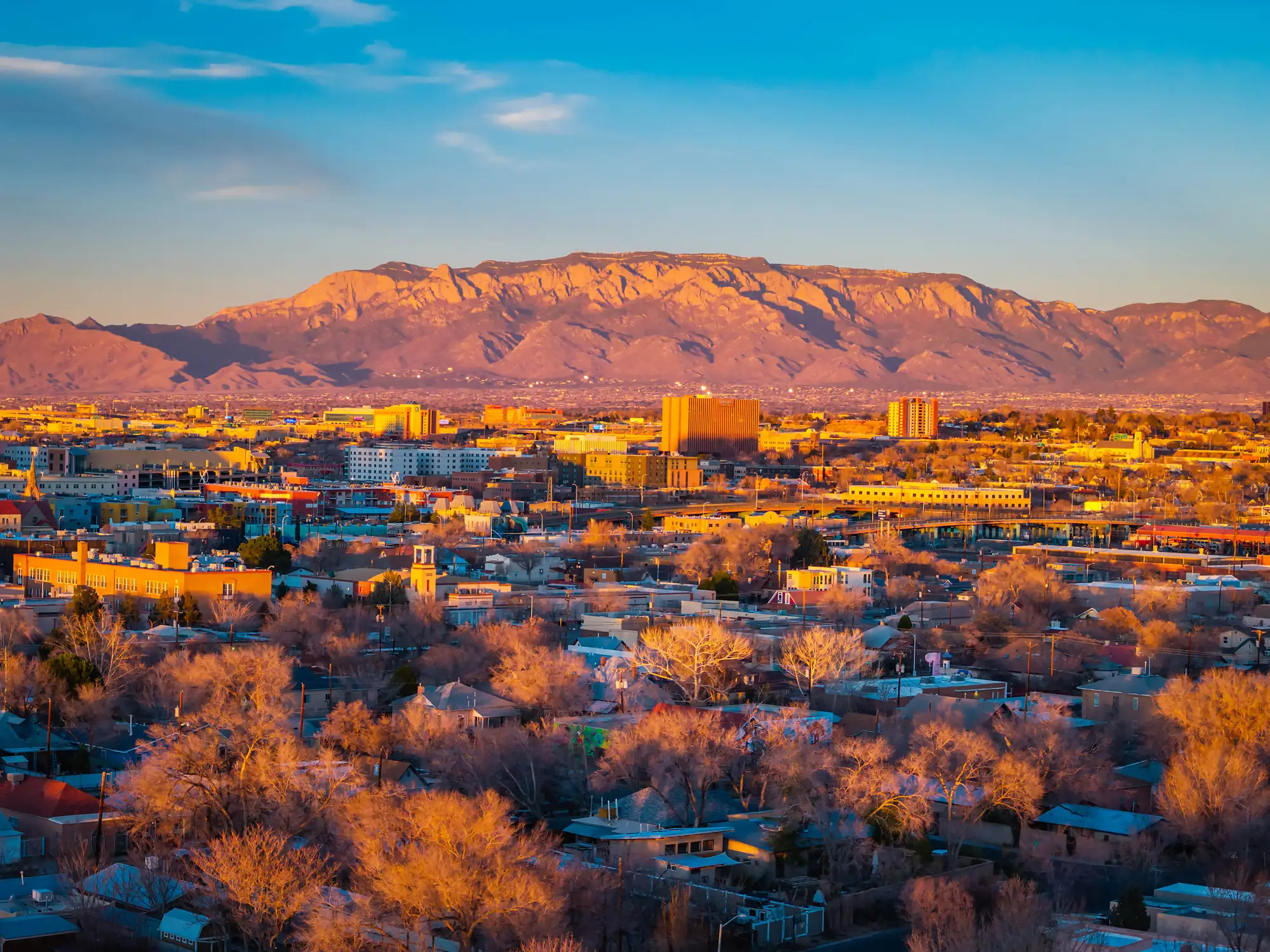 Albuquerque cityscape at golden hour with the Sandia Mountains rising behind downtown buildings and residential neighborhoods.