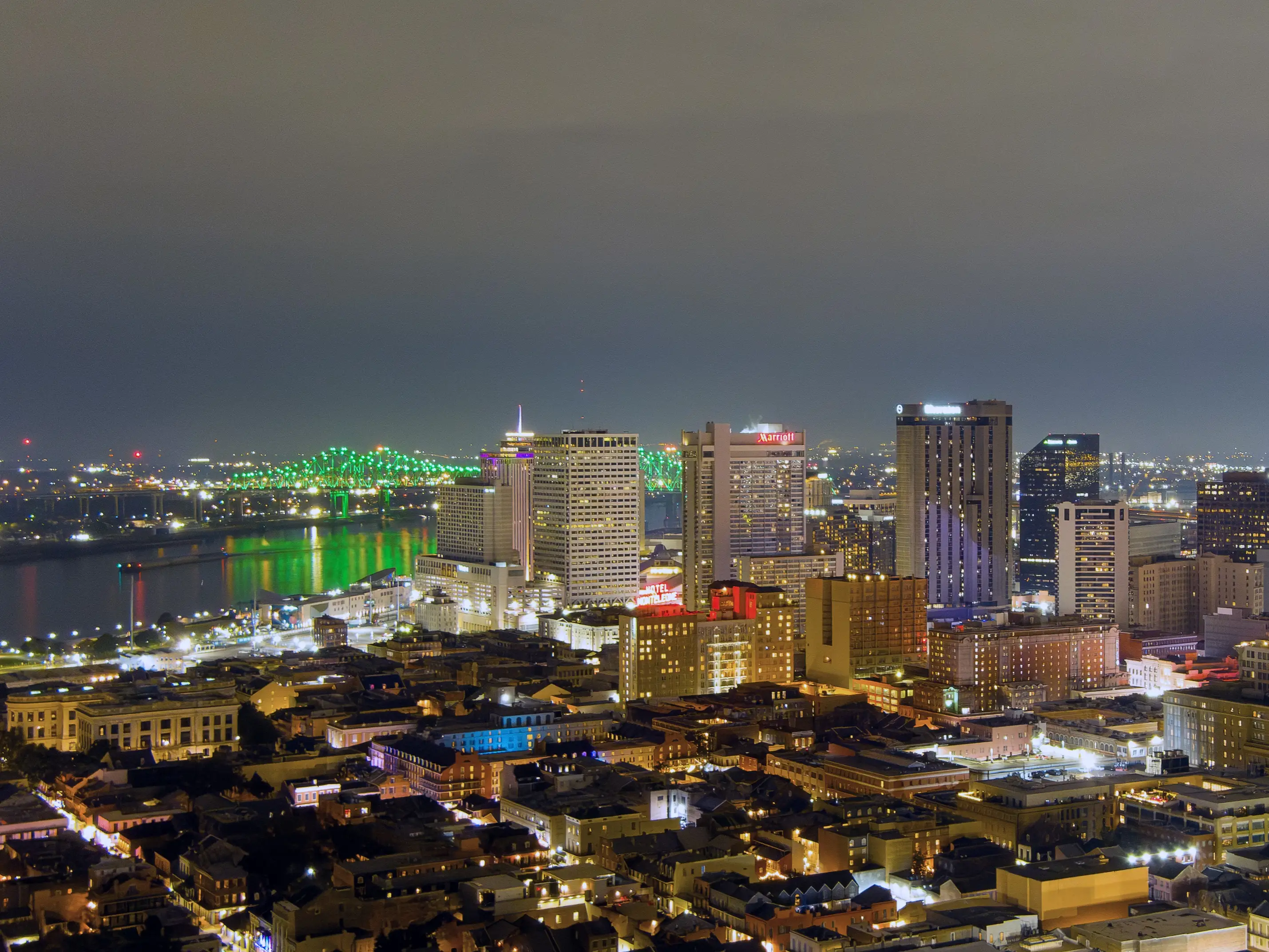 Nighttime New Orleans skyline with illuminated high-rises and a green-lit bridge over the Mississippi River.