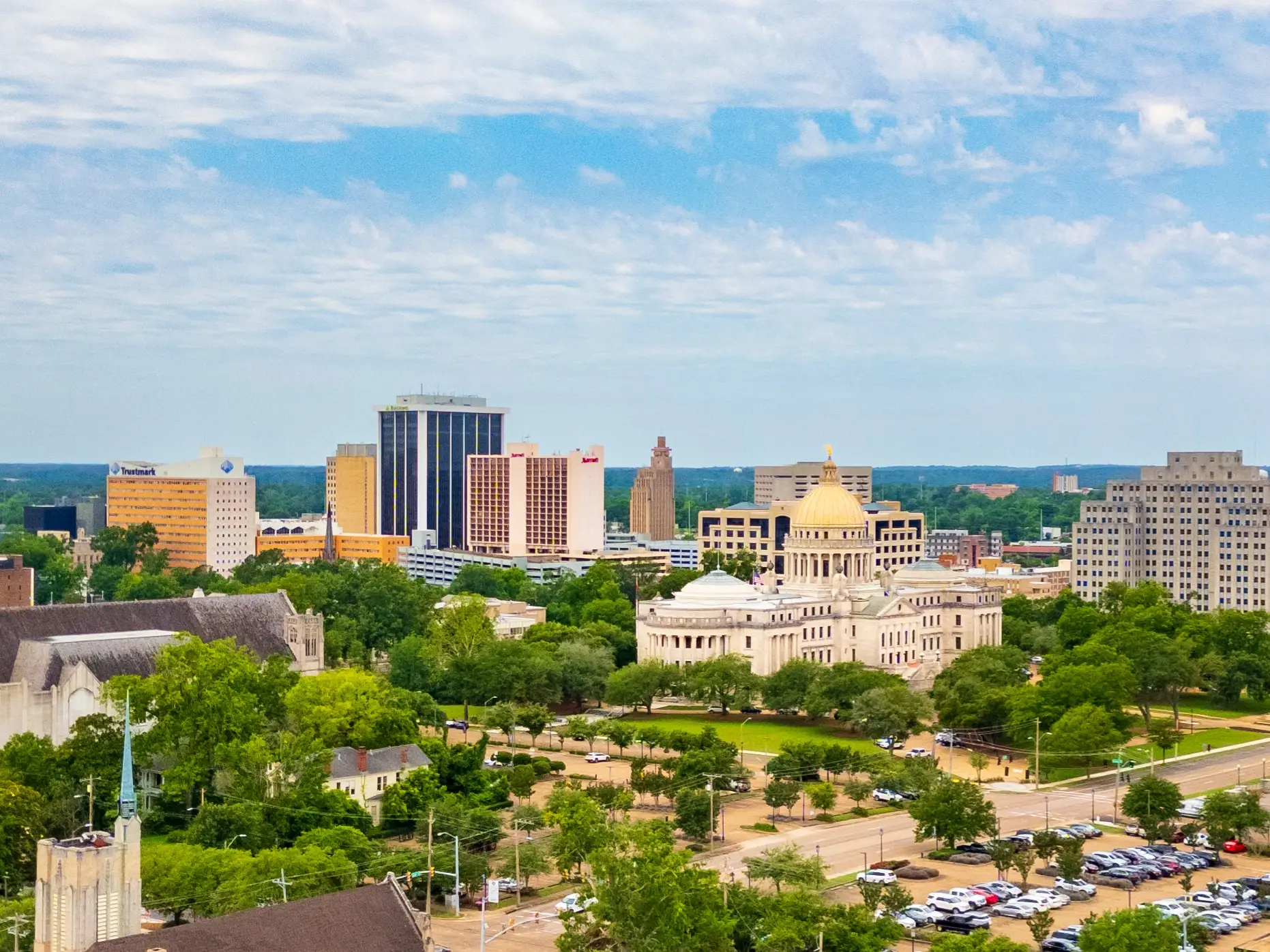 Downtown Jackson skyline with the Mississippi State Capitol’s gold dome, office towers, trees, roads, and parking lots.
