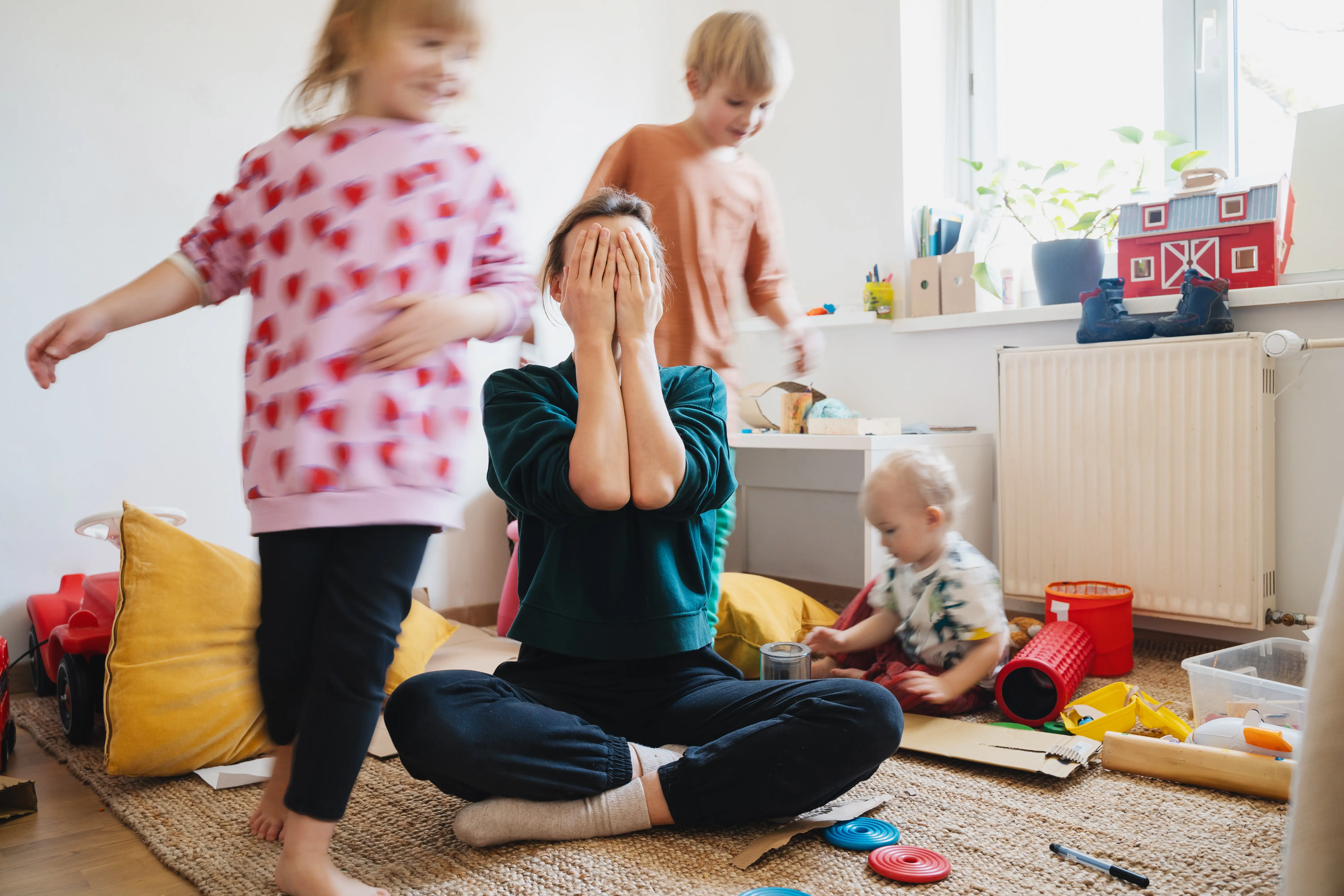 A mom holds her hands over her face while children run around her.