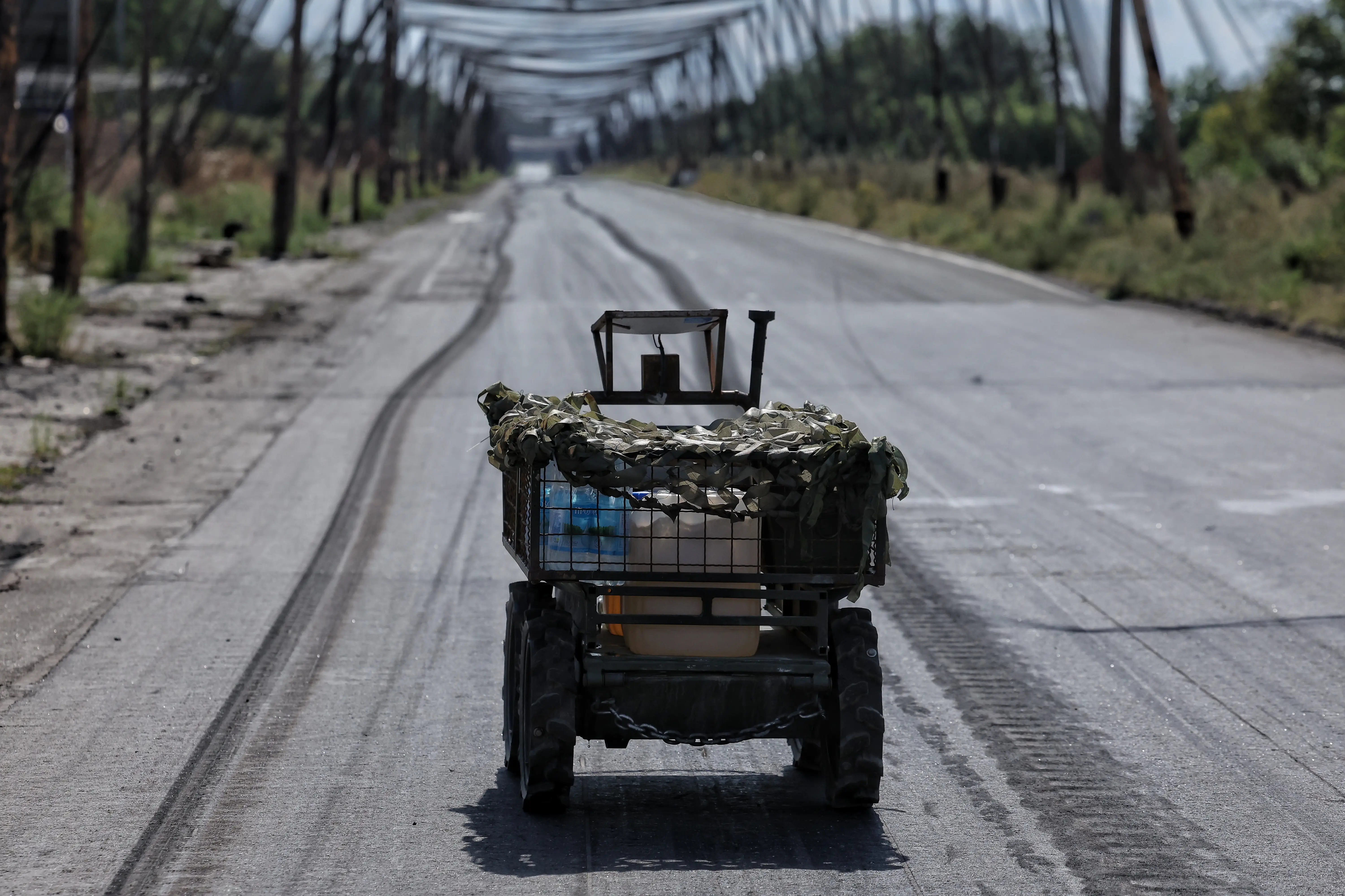 A small camouflaged four-wheeled vehicle sits on an empty road beneath a metal bridge structure.