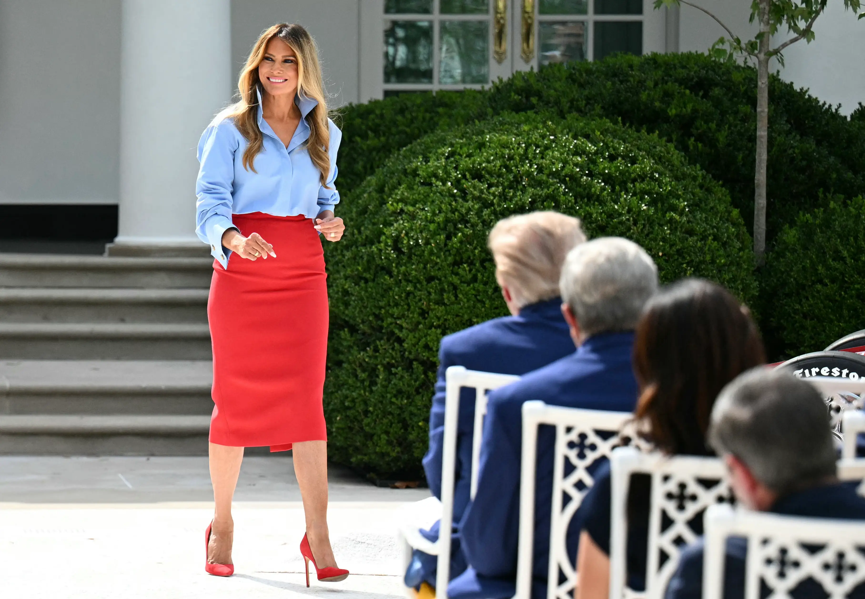Melania Trump stands outdoors beside seated attendees near White House columns and greenery.