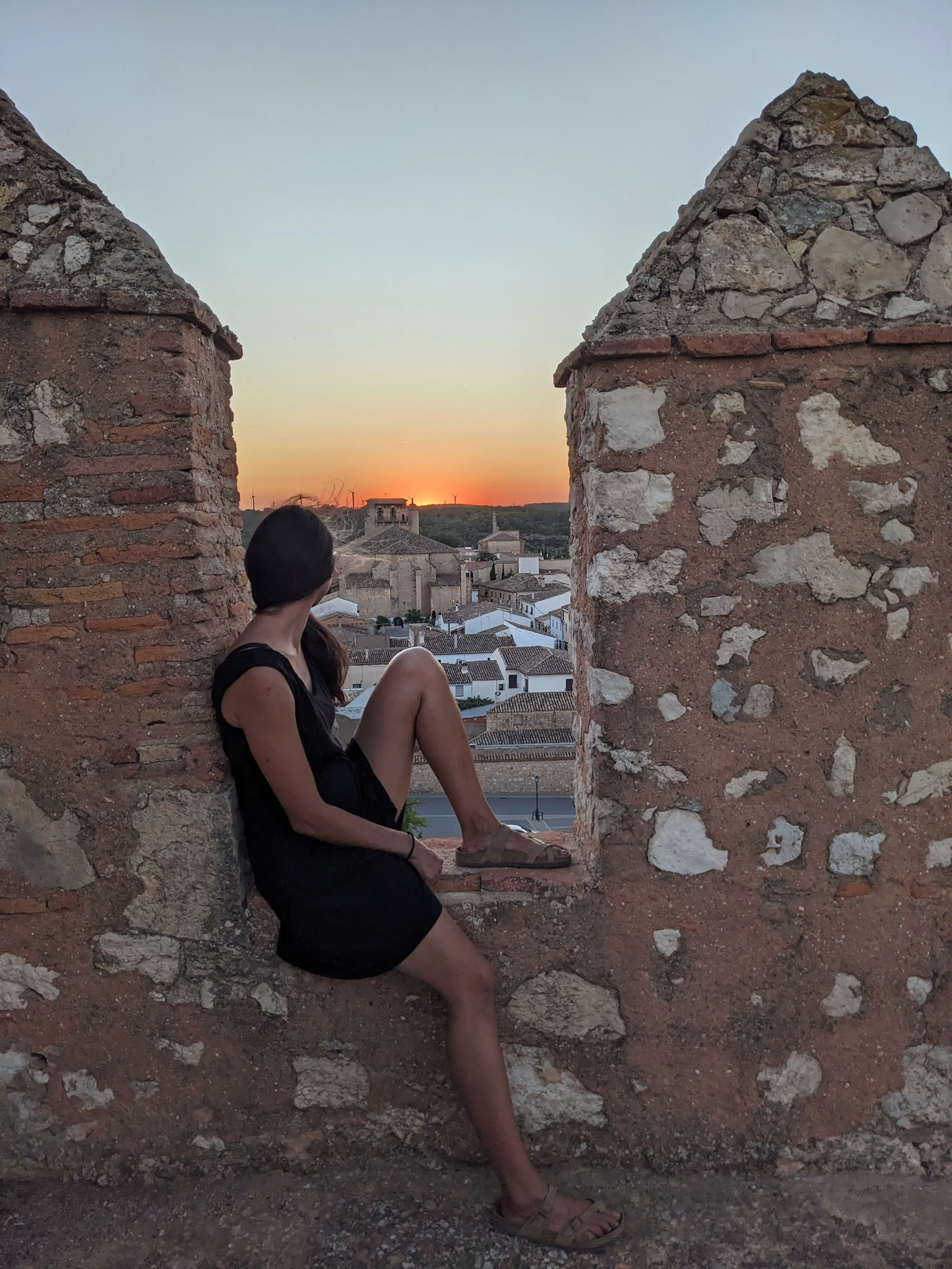 A seated person in a black dress looks over rooftops from an old stone wall at sunset.