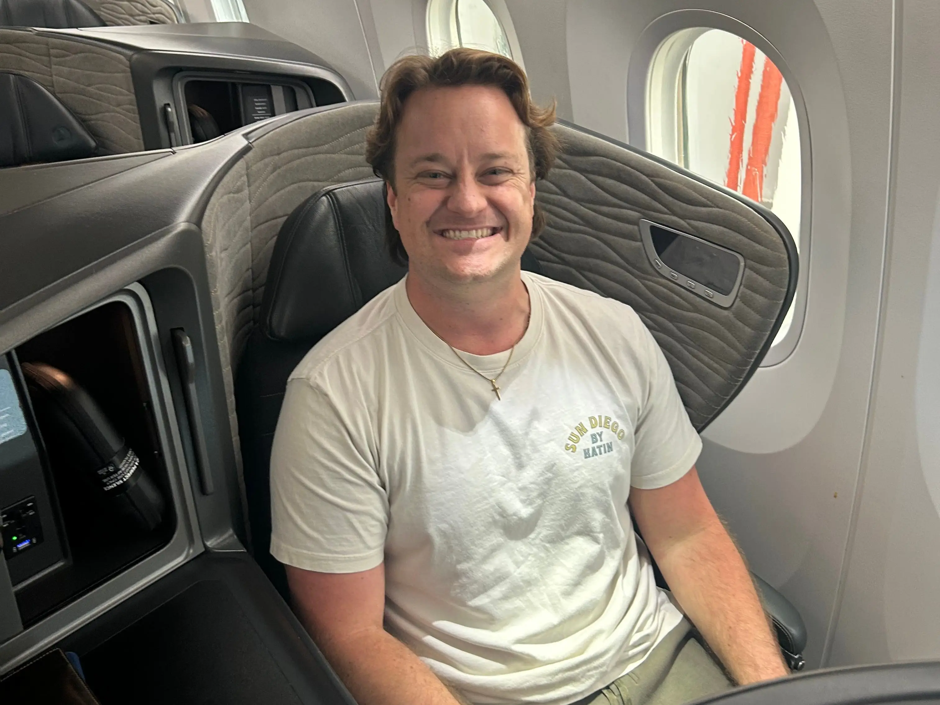Smiling passenger seated in an airplane cabin seat beside oval windows.