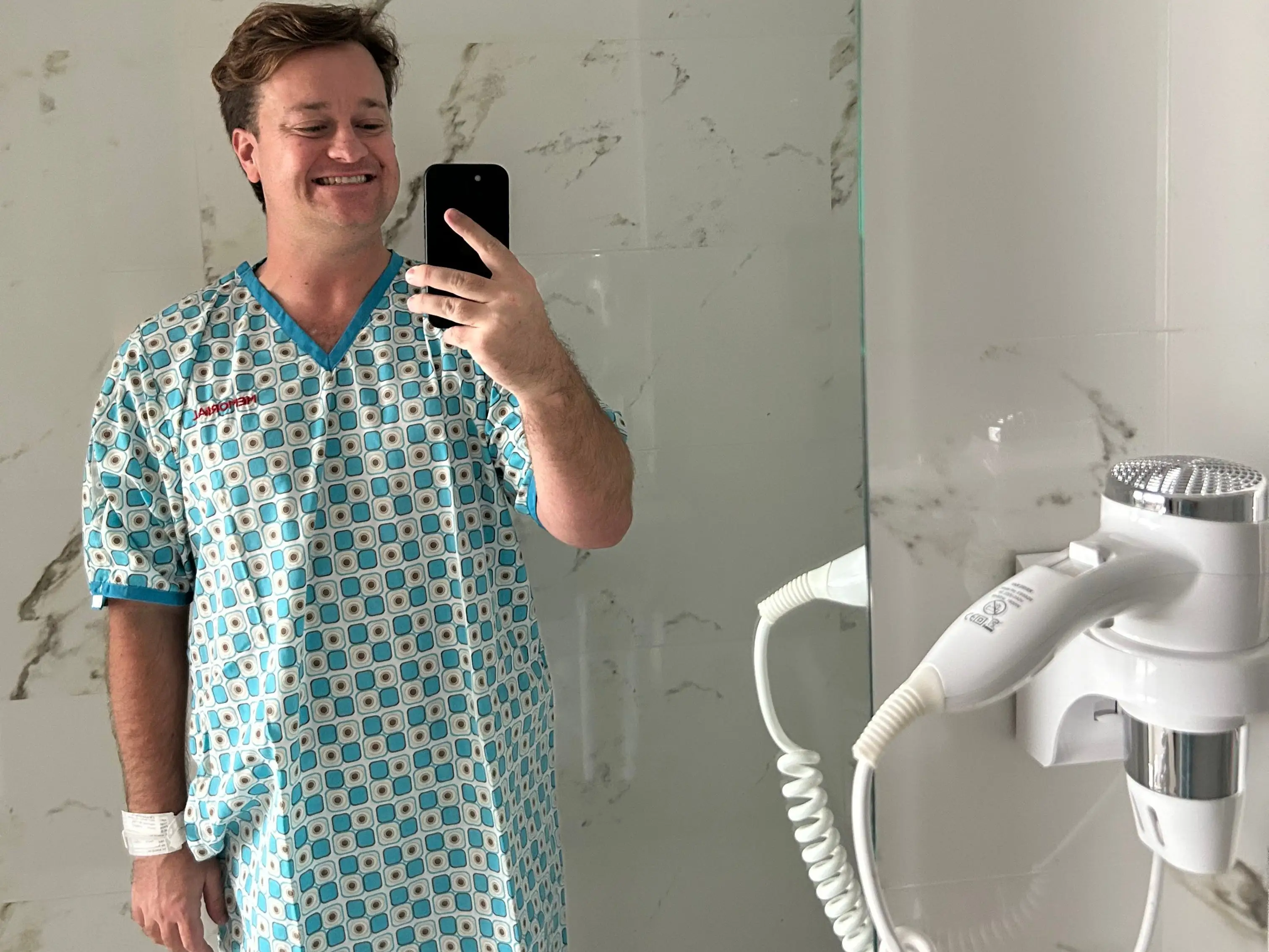 A smiling person in a patterned hospital gown takes a mirror selfie in a bathroom with marble walls.