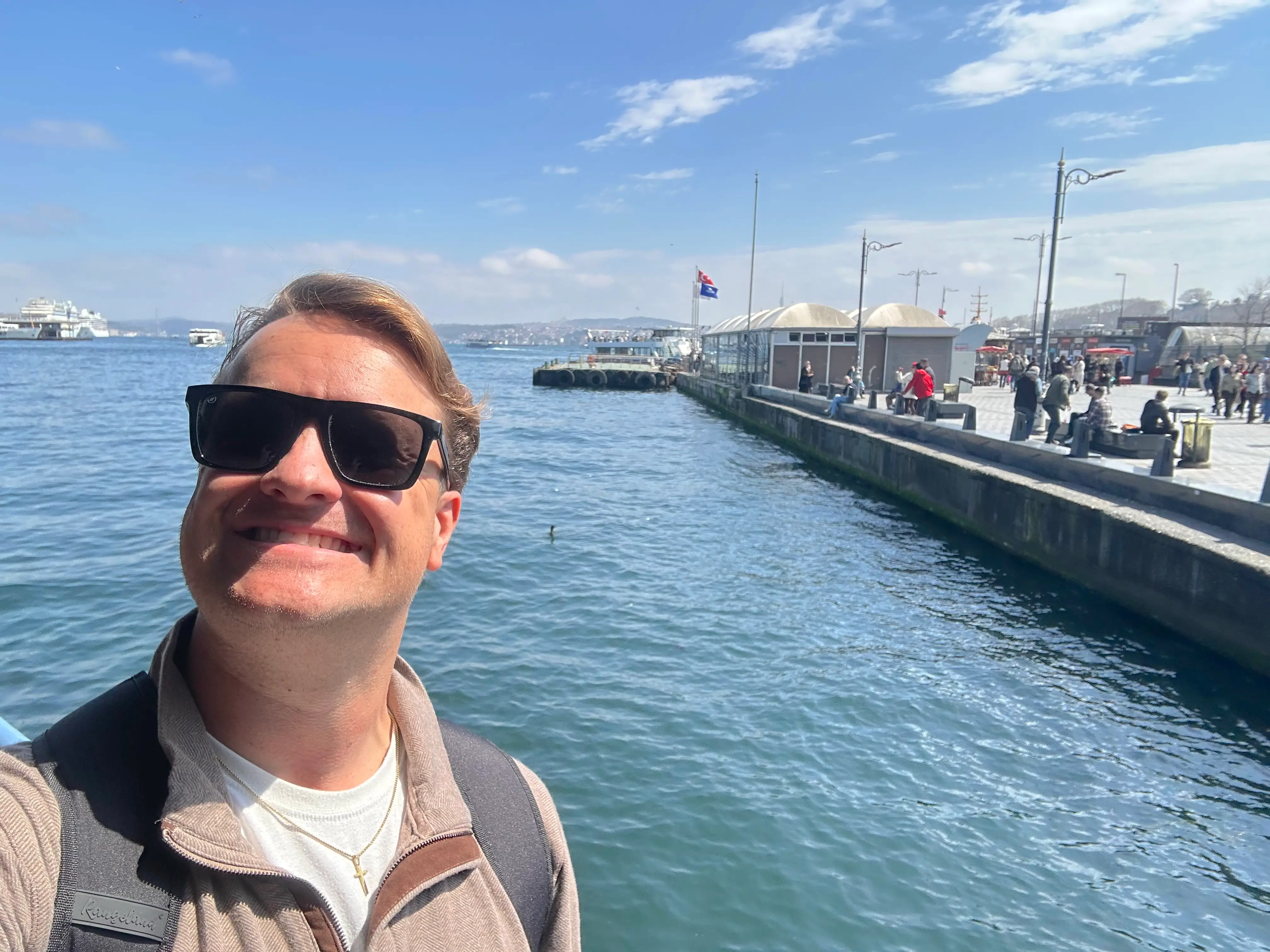 Person wearing sunglasses takes a waterfront selfie beside a pier with boats and pedestrians under a blue sky.