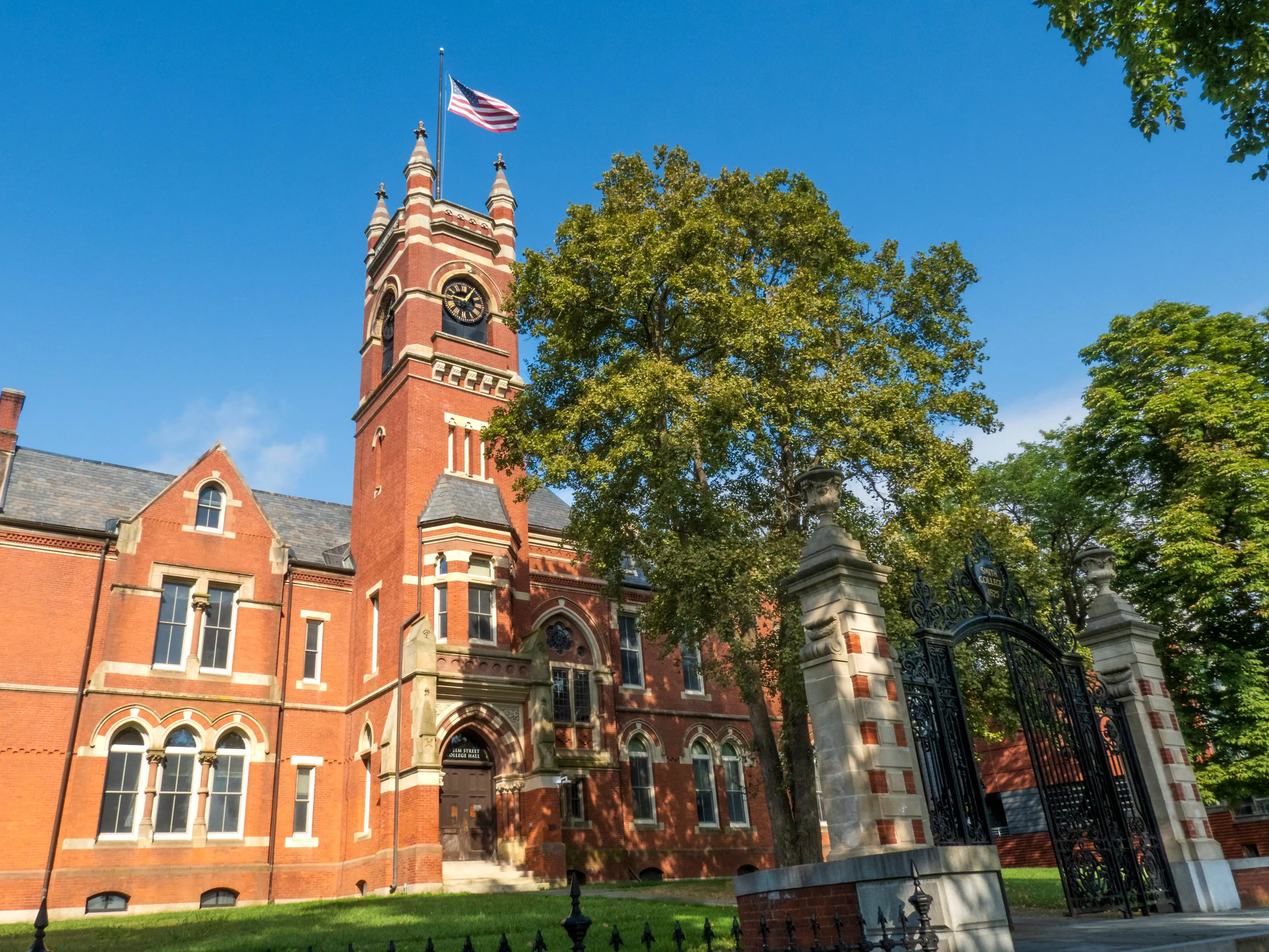 Northampton, MA - US - Aug 24, 2025 A grand brick College Hall at Smith College, with a prominent clock tower and American flag, set against a clear blue sky amidst lush green trees.