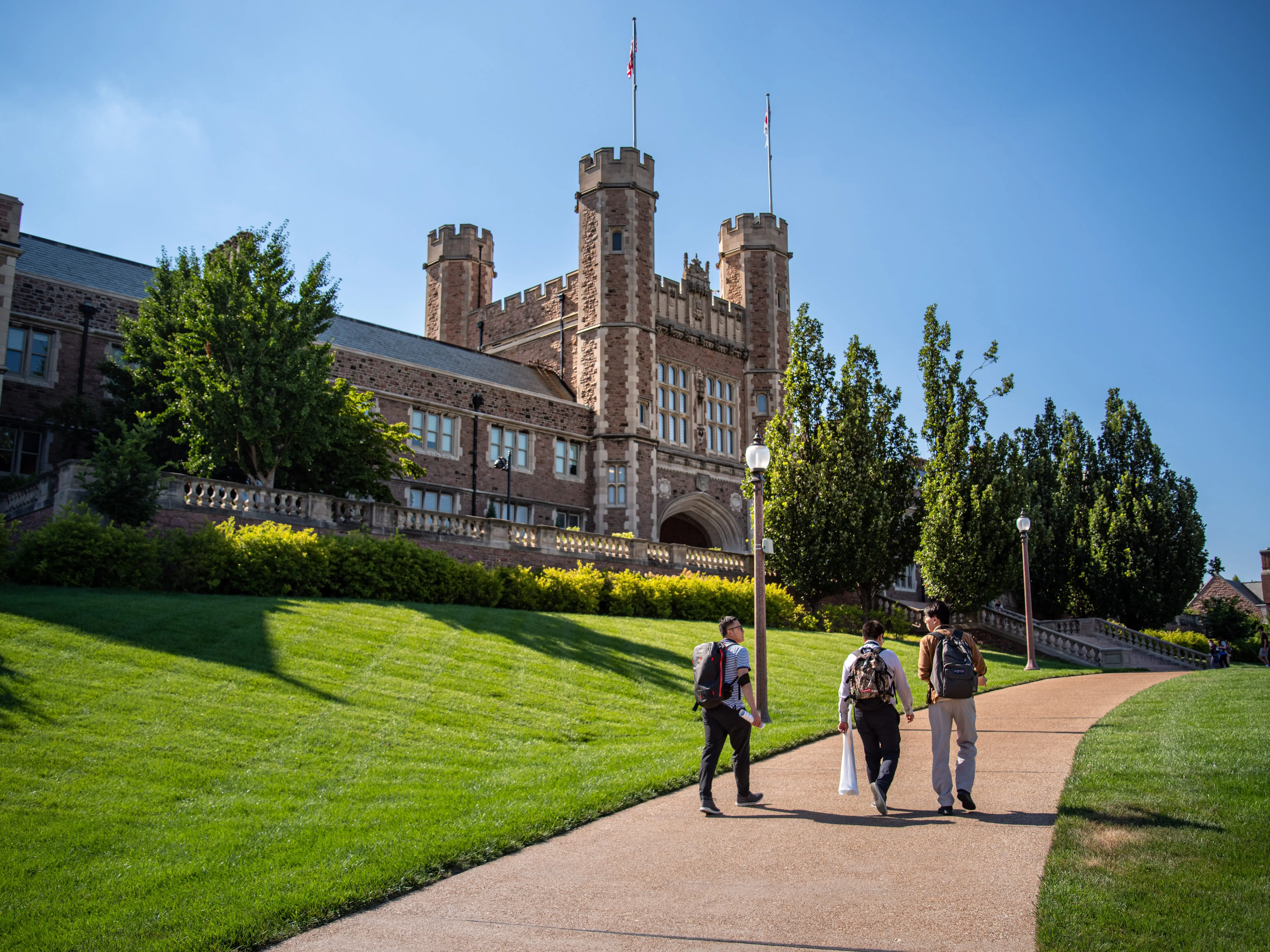 Three people with backpacks on sidewalk in front of the campus administrative building on sunny day moving away.
