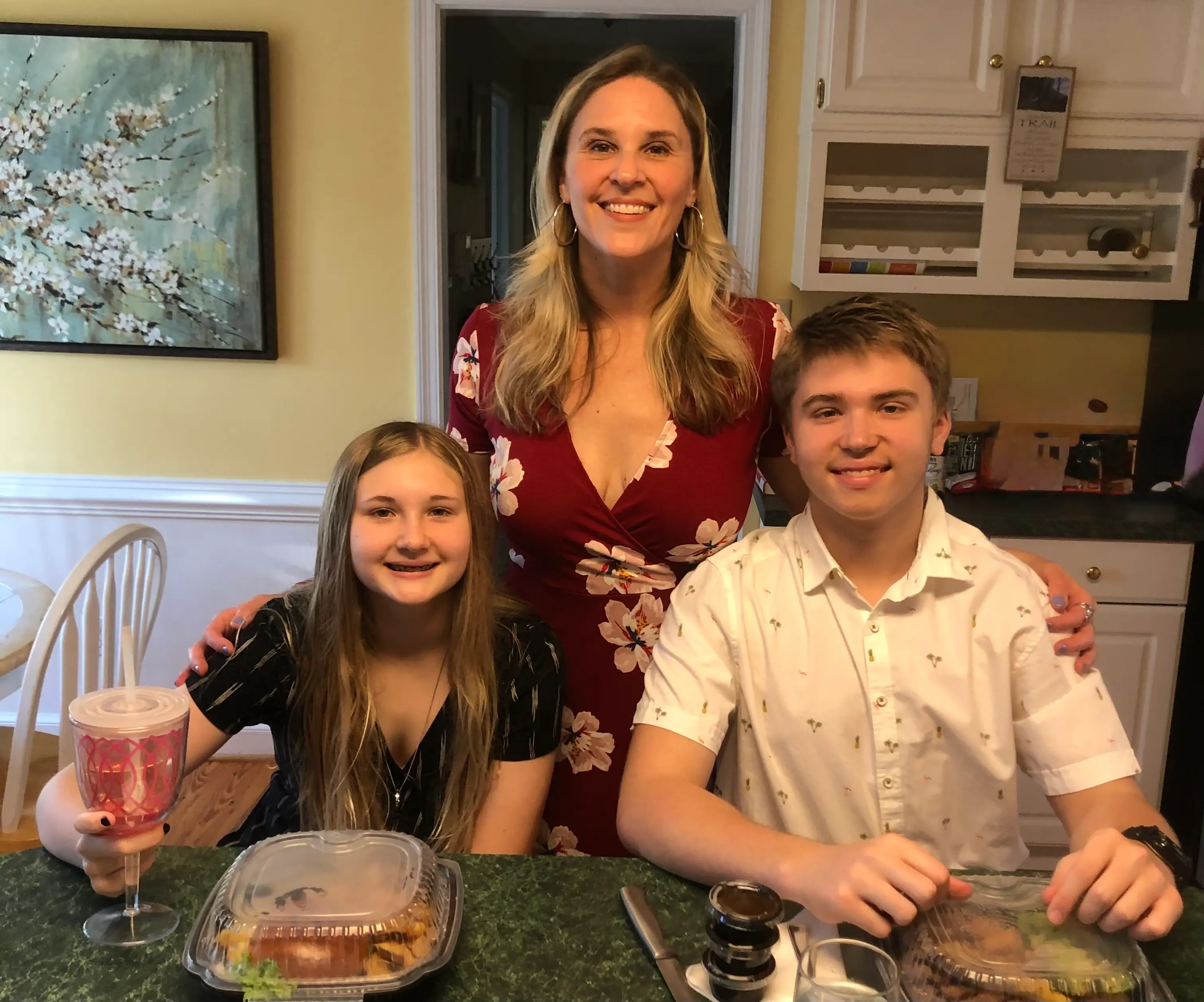 Three smiling people pose at a kitchen counter with takeout containers, cups, and cabinets behind them.