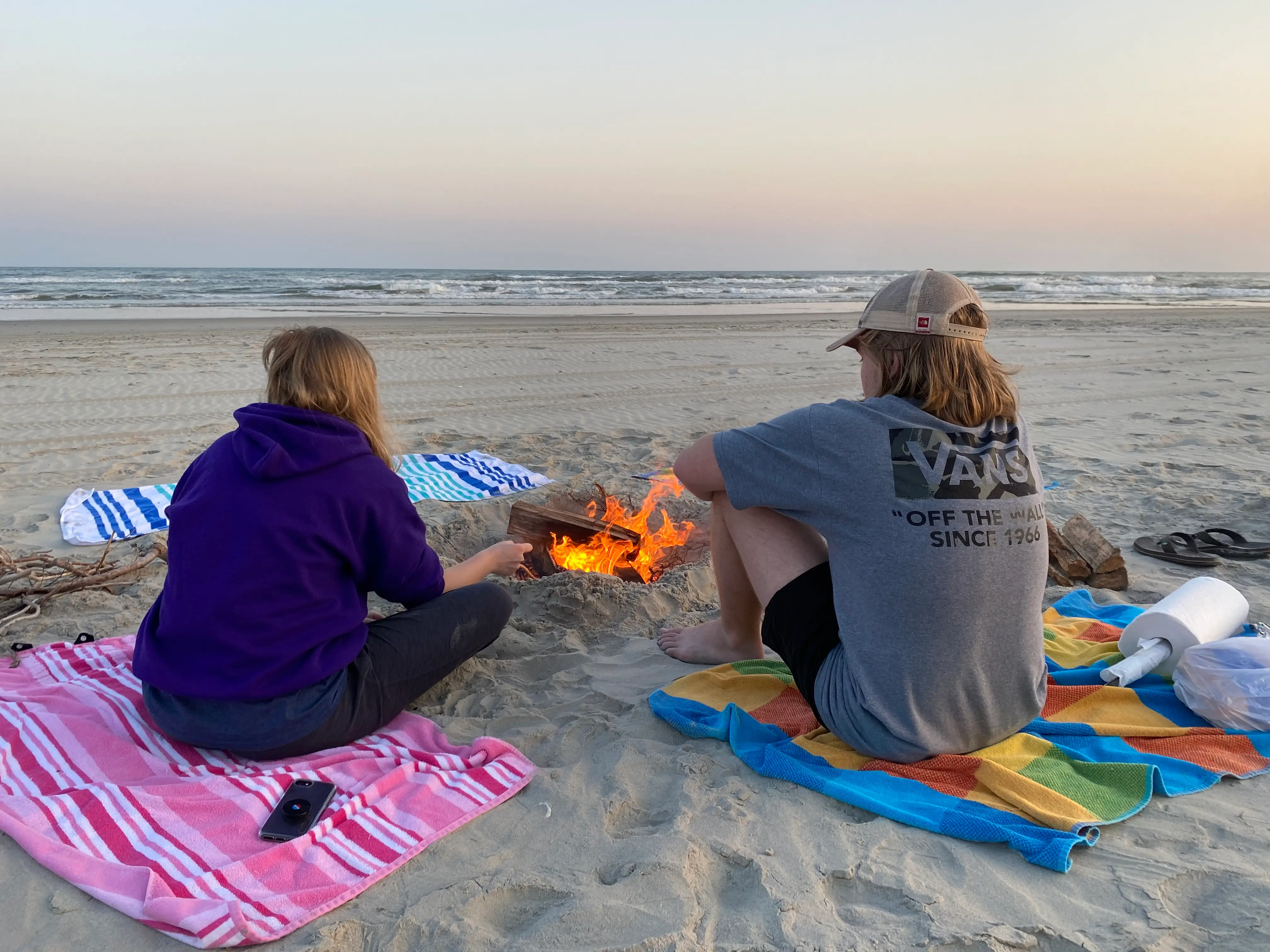 Two people sit on beach towels beside a small fire on the sand with ocean waves in the background.