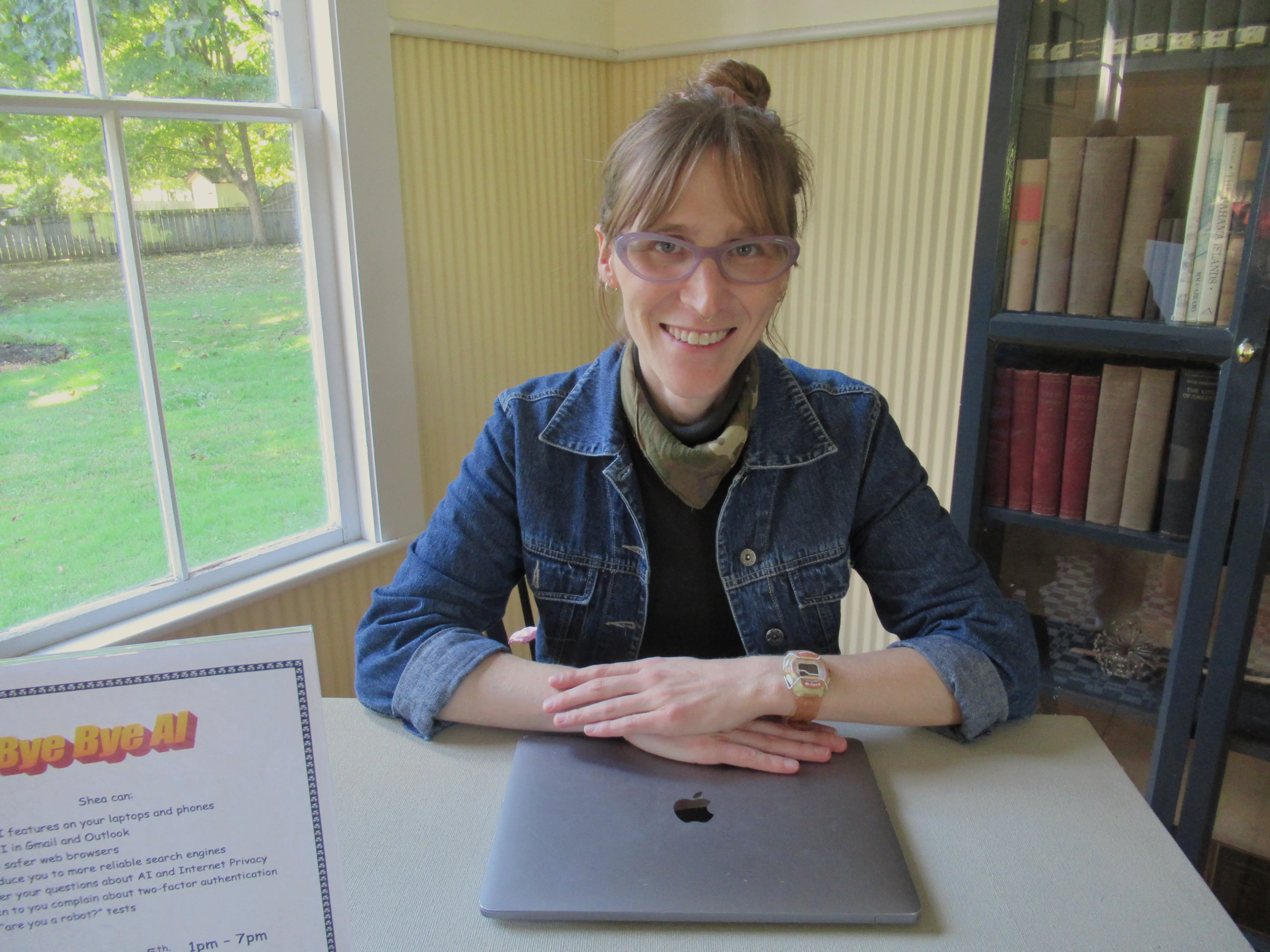 Smiling person with purple glasses sits at a table with an Apple laptop beside a “Bye Bye AI” sign.
