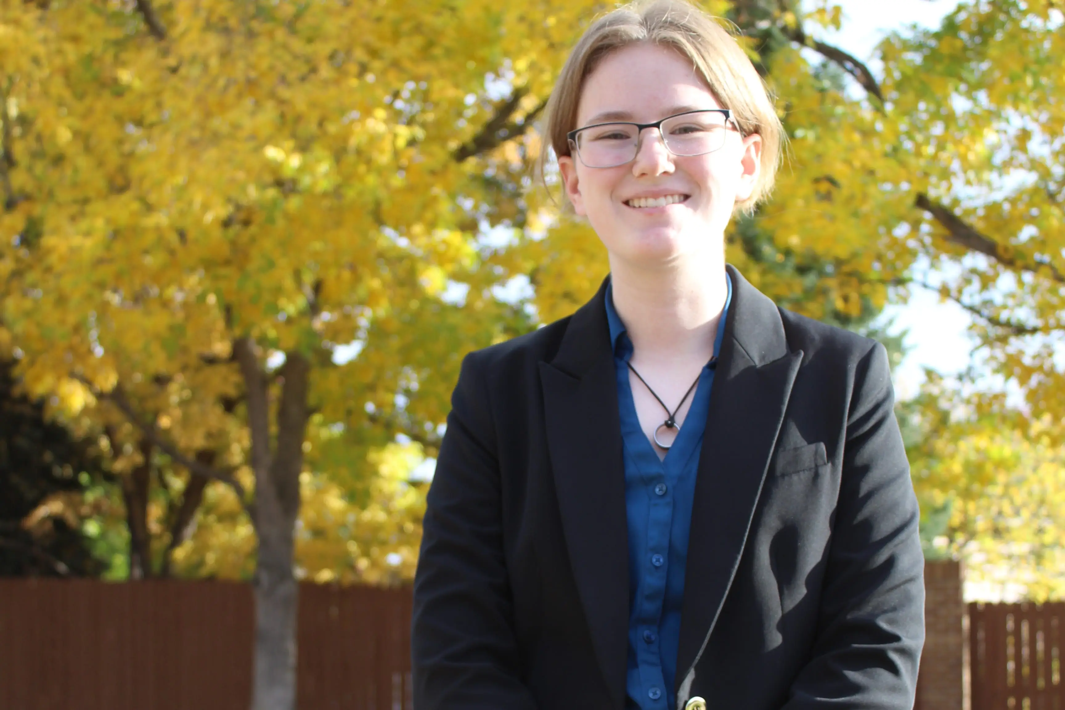 Hannah Mitchell is in a black blazer and a blue button-up, standing in front of trees with beautiful yellow fall leaves.