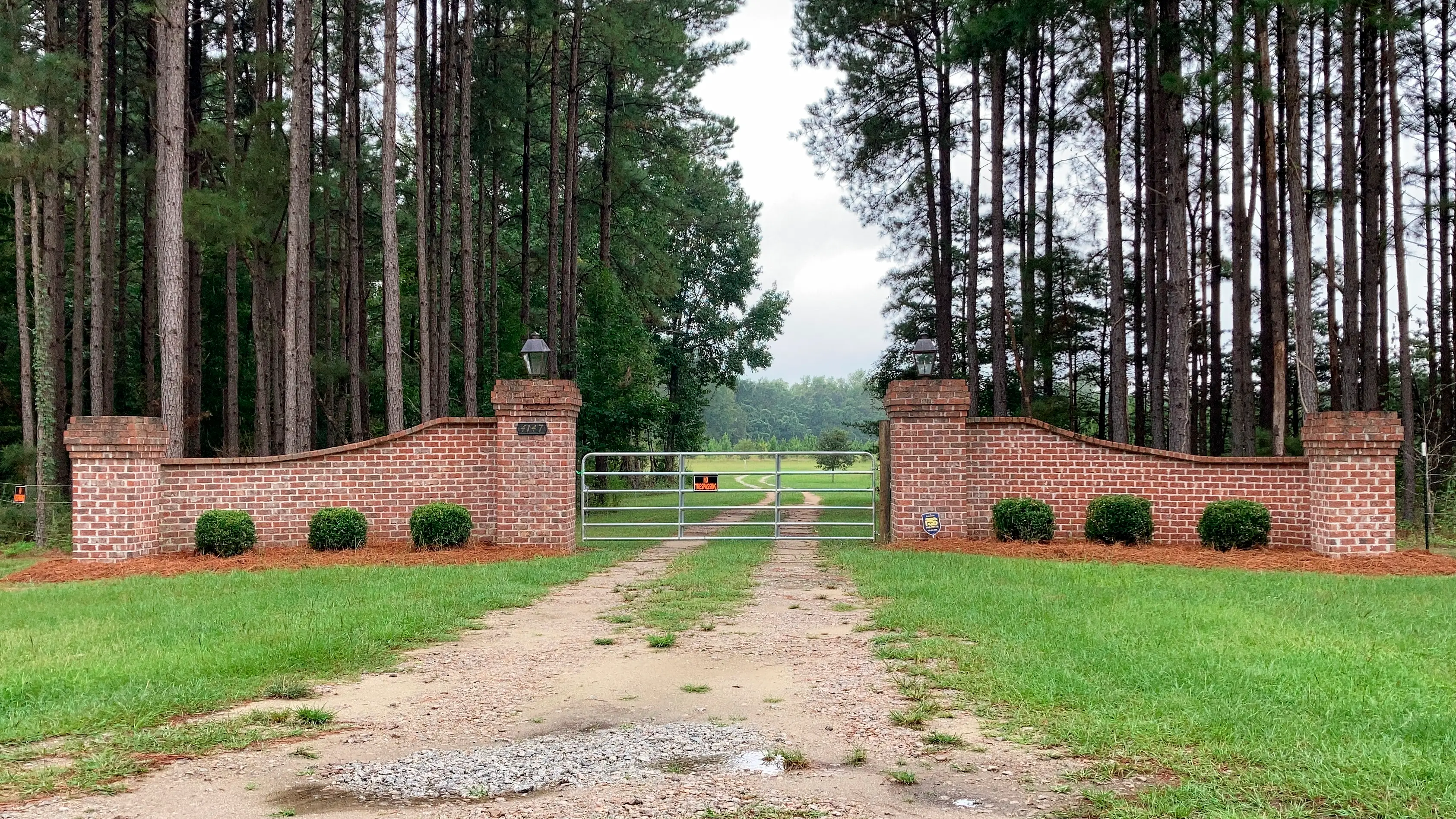 A brick-walled, gated driveway entrance stands between tall pine trees, with a gravel track and a green lawn.