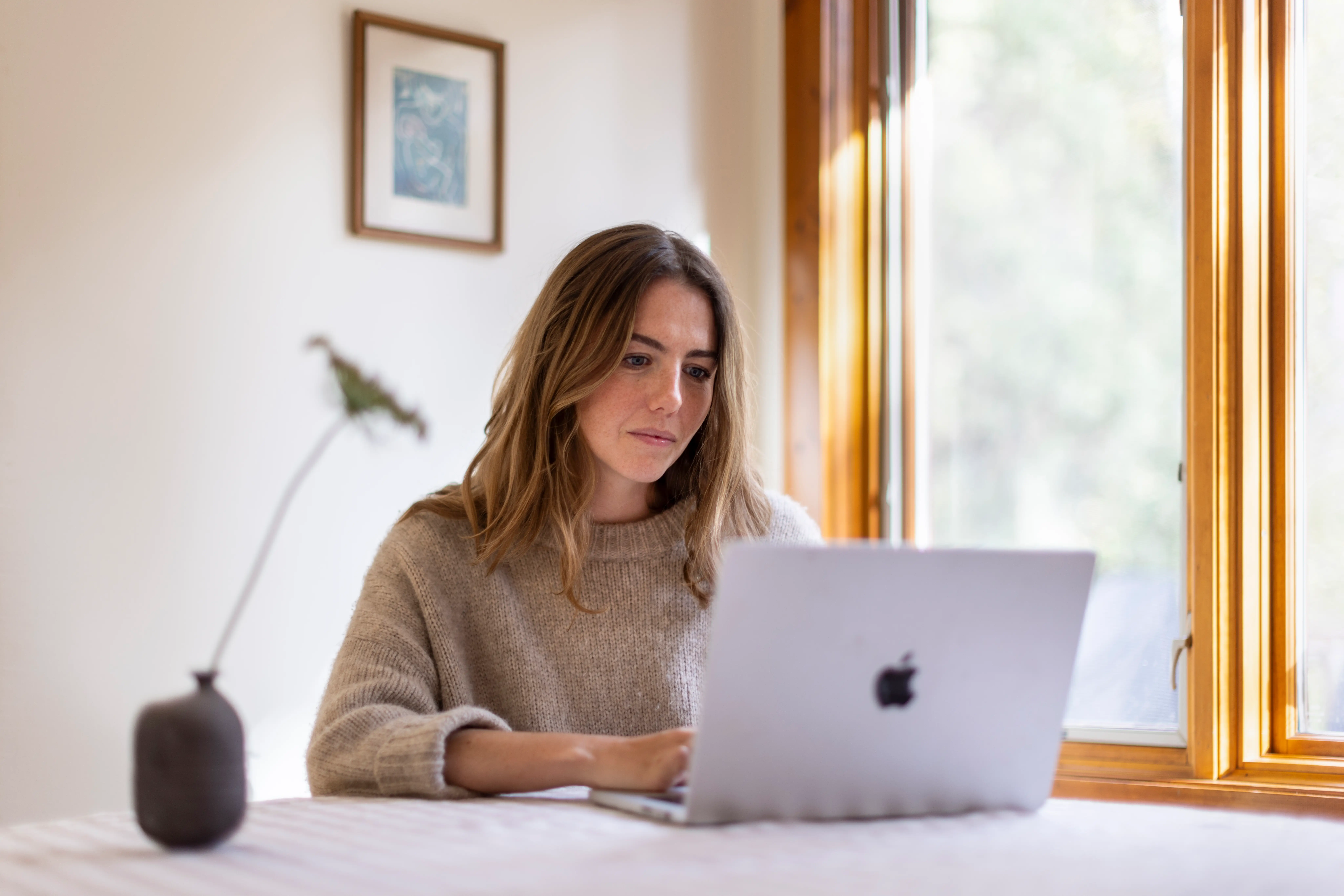 Emily Byrne works on an Apple laptop beside a sunlit wooden-framed window.