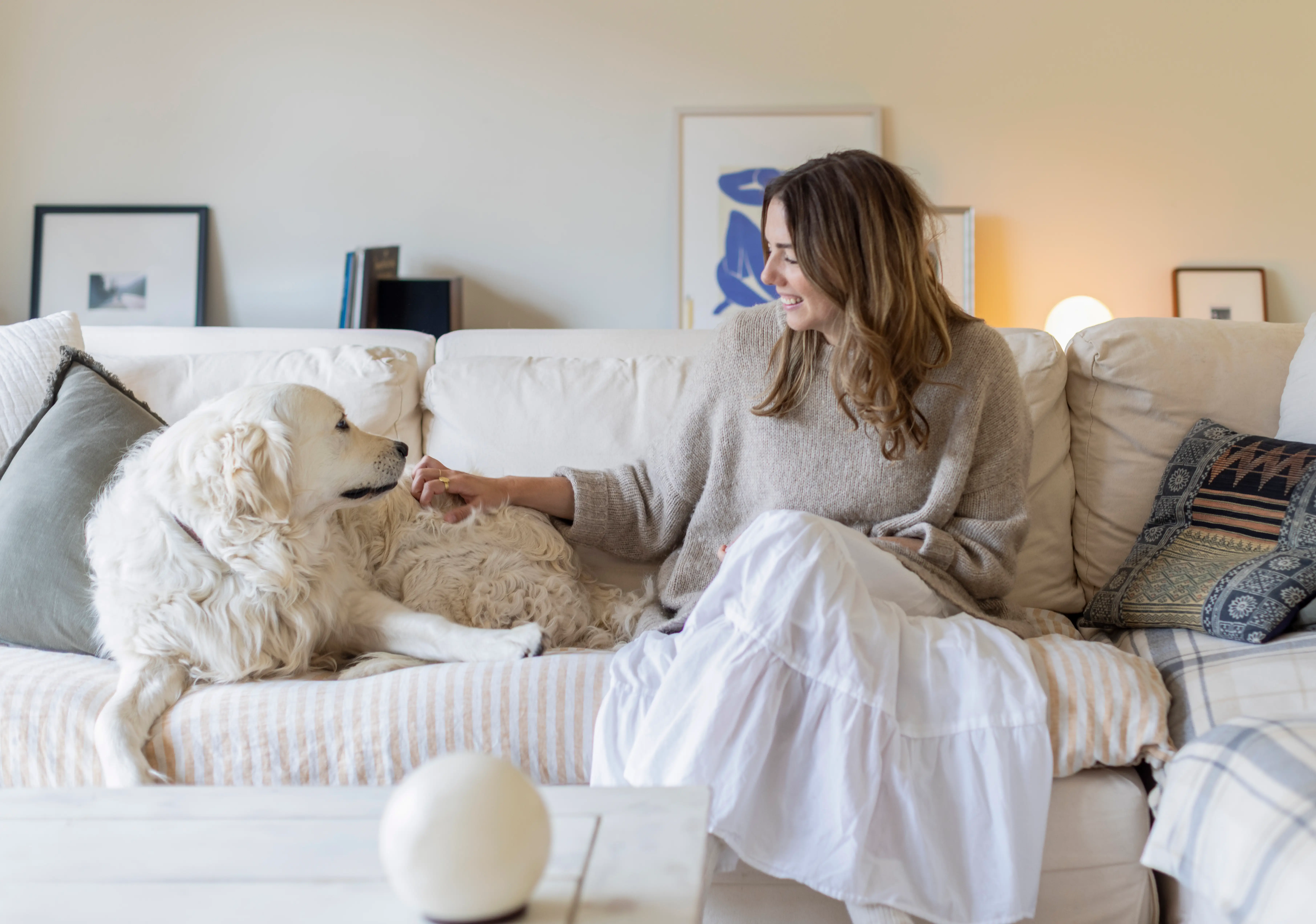 Emily Byrne sits on a cream sofa and pets a large white dog in a softly lit living room.