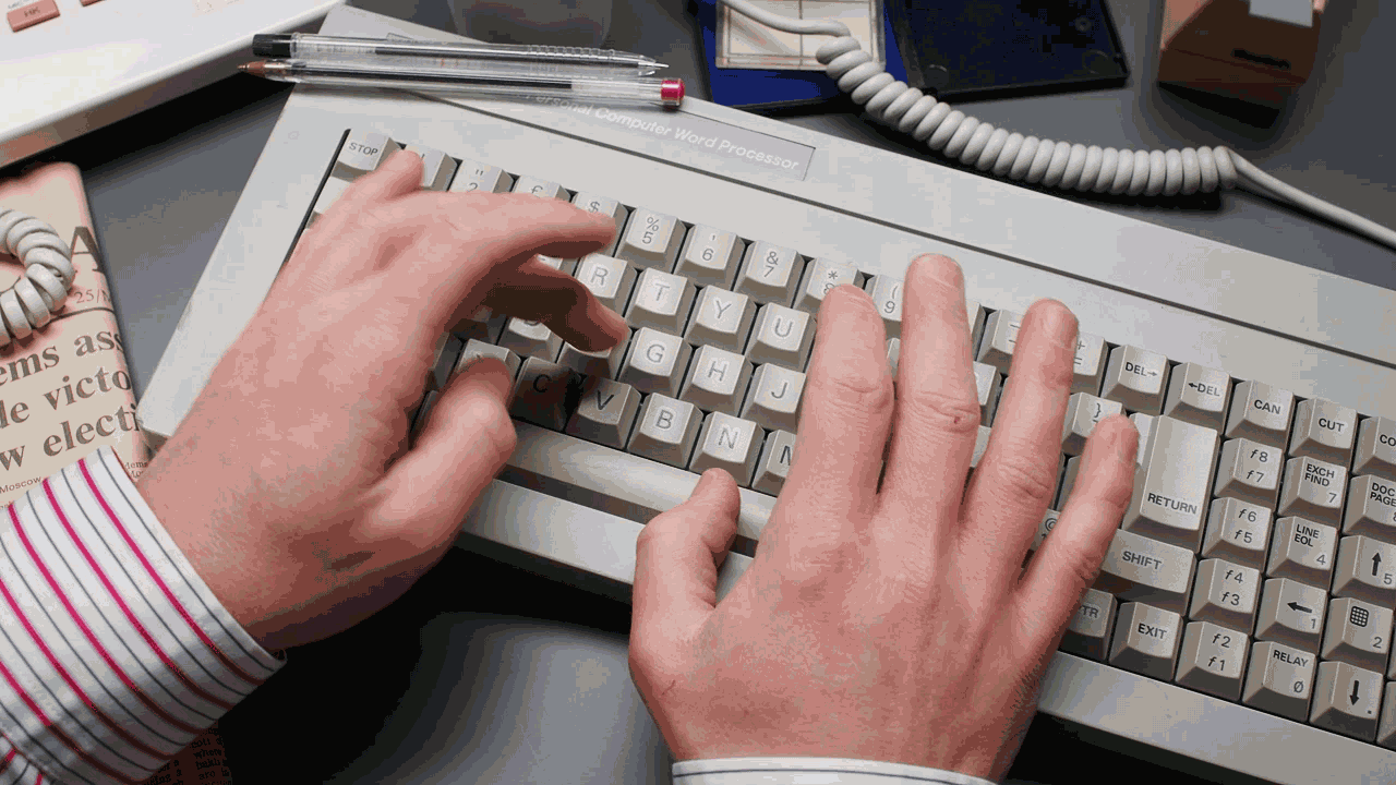 Male hands with pinstripe shirt cuffs typing on a retro 1980s word processor keyboard.