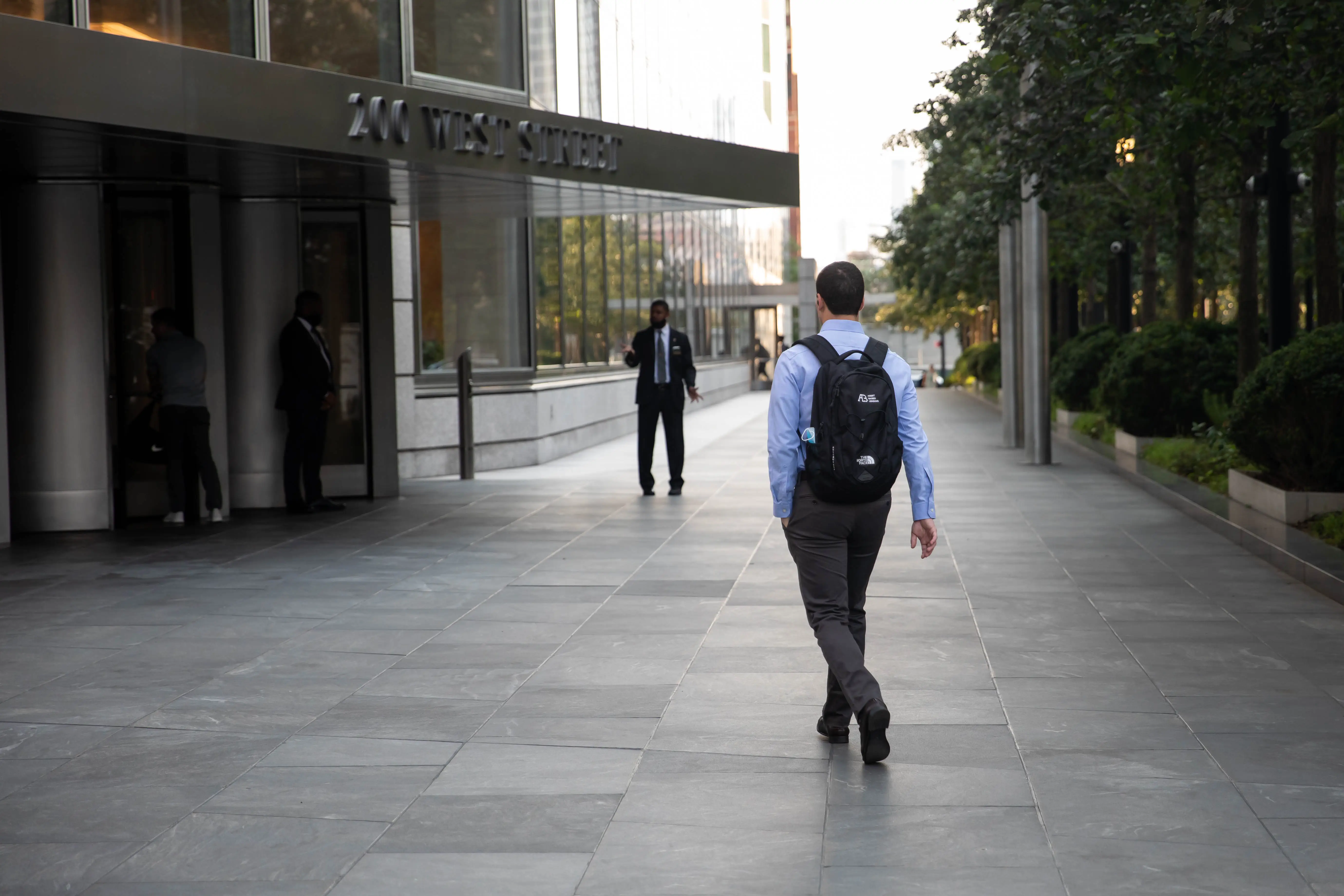 A person with a backpack walks toward a modern office tower entrance along a tree-lined paved walkway.