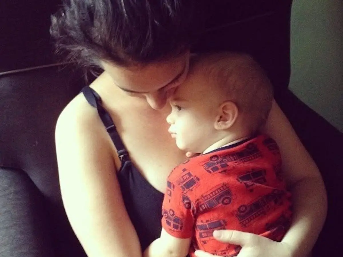 Adult cuddles a baby in a red patterned shirt while seated on a dark couch.