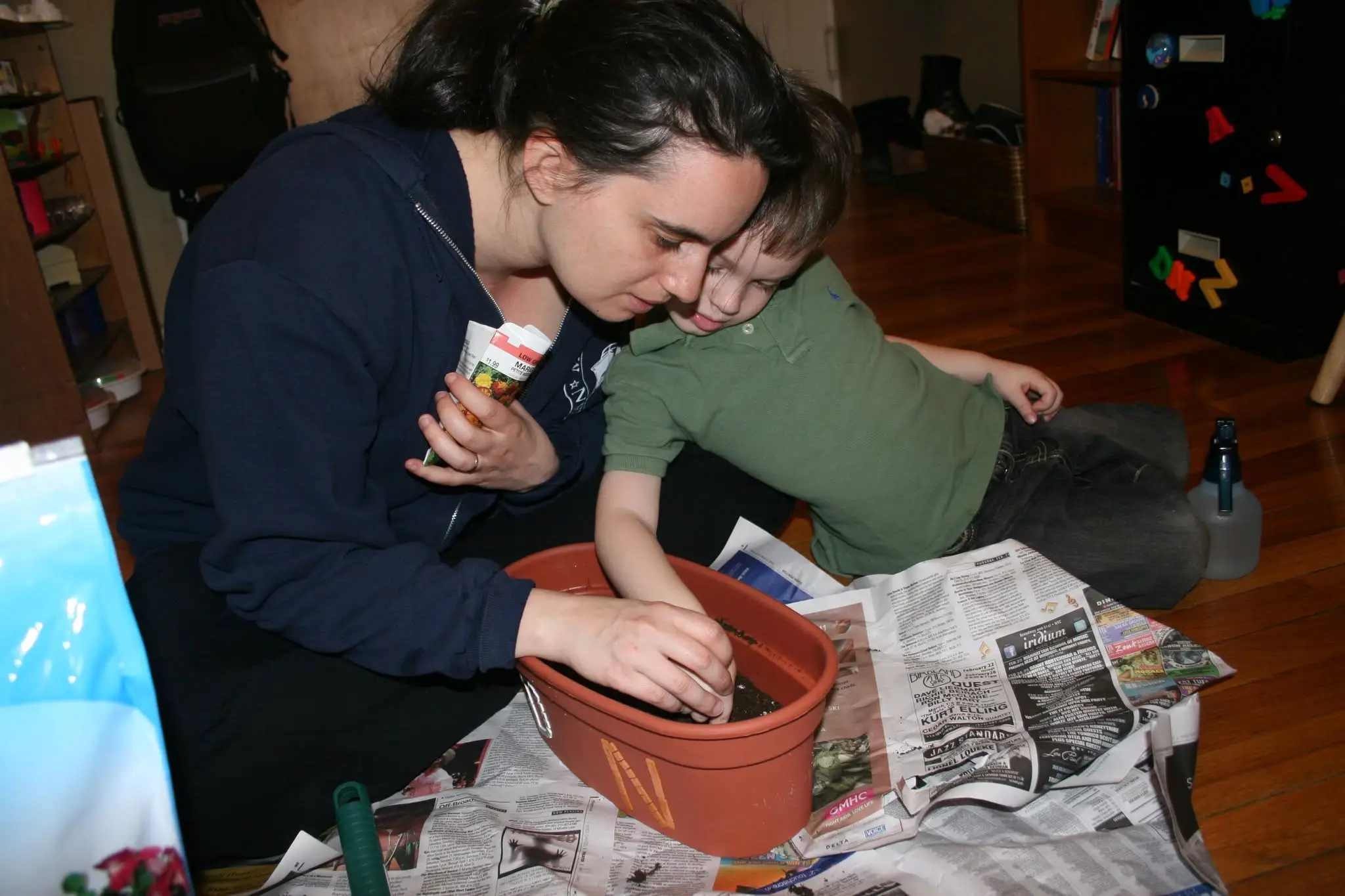 Two people lean over a terracotta planter filled with soil on newspapers indoors.
