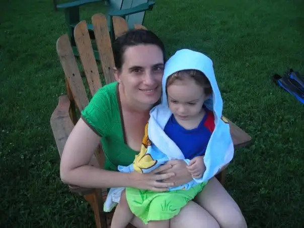 Person seated on a wooden lawn chair holding a small child wrapped in a hooded towel on grass.