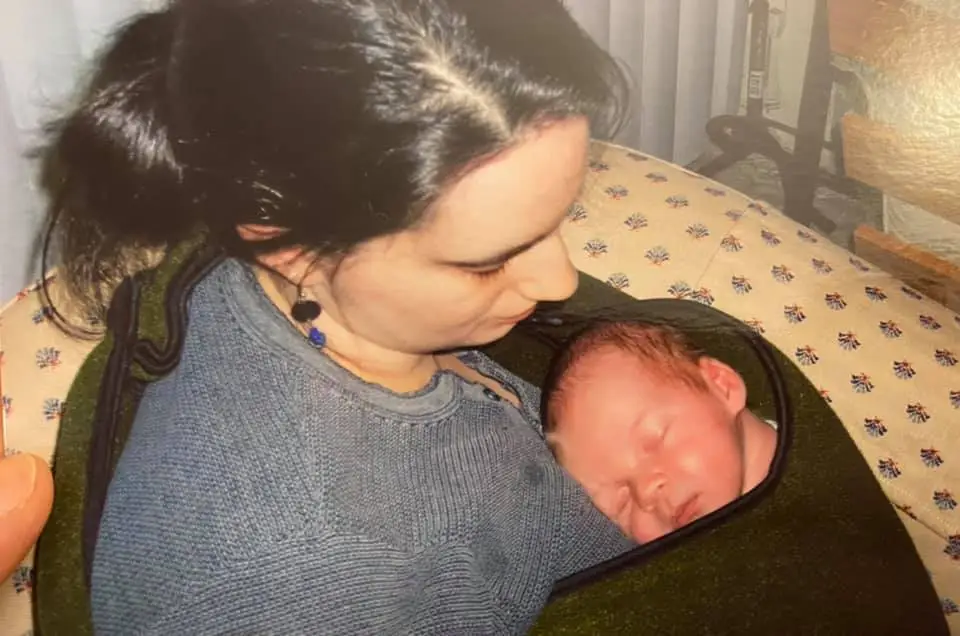 A person in a gray sweater holds a sleeping baby wrapped in a dark blanket indoors.