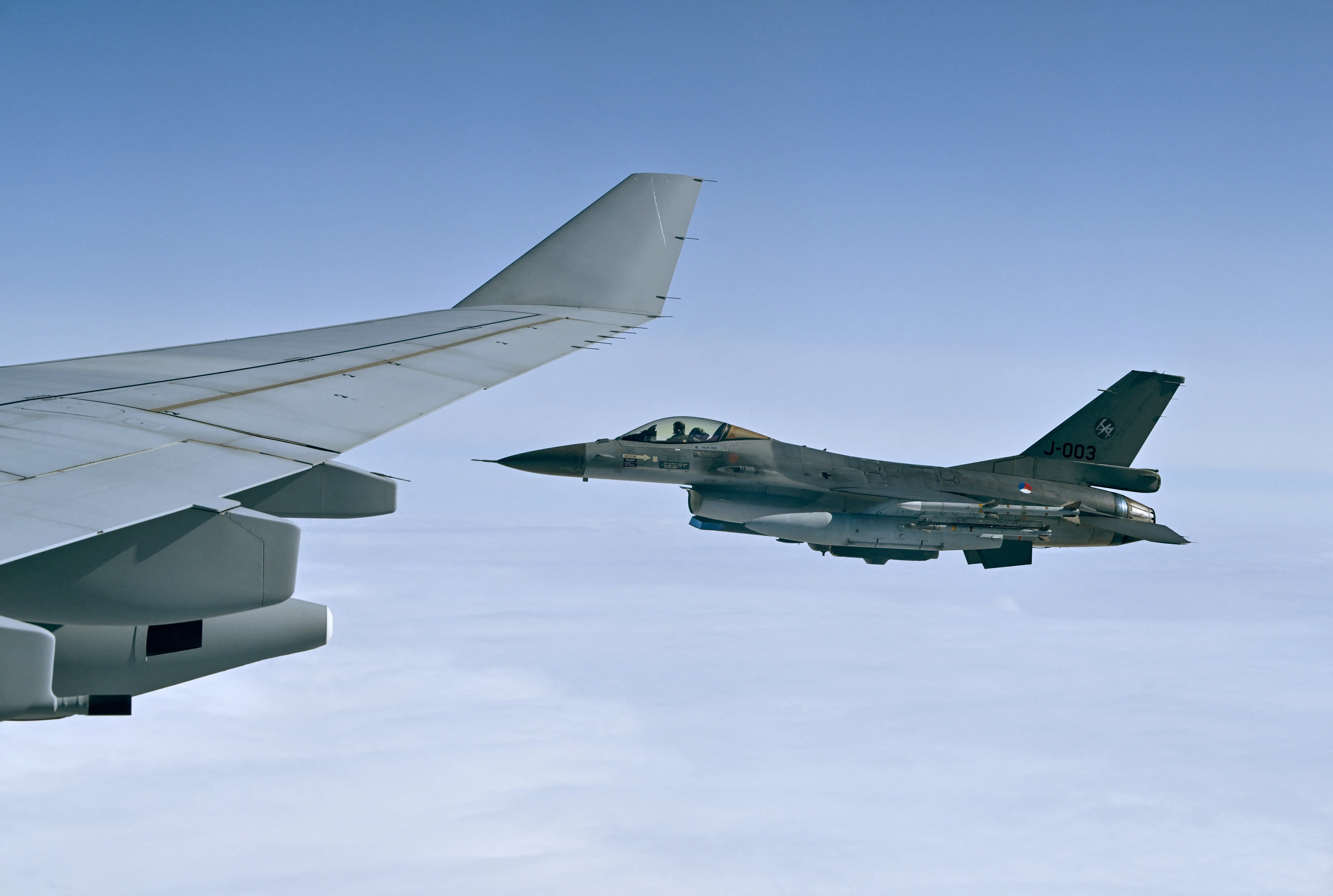 F-16 fighter jet marked J-009 flies beside a large aircraft wing above a cloud layer.