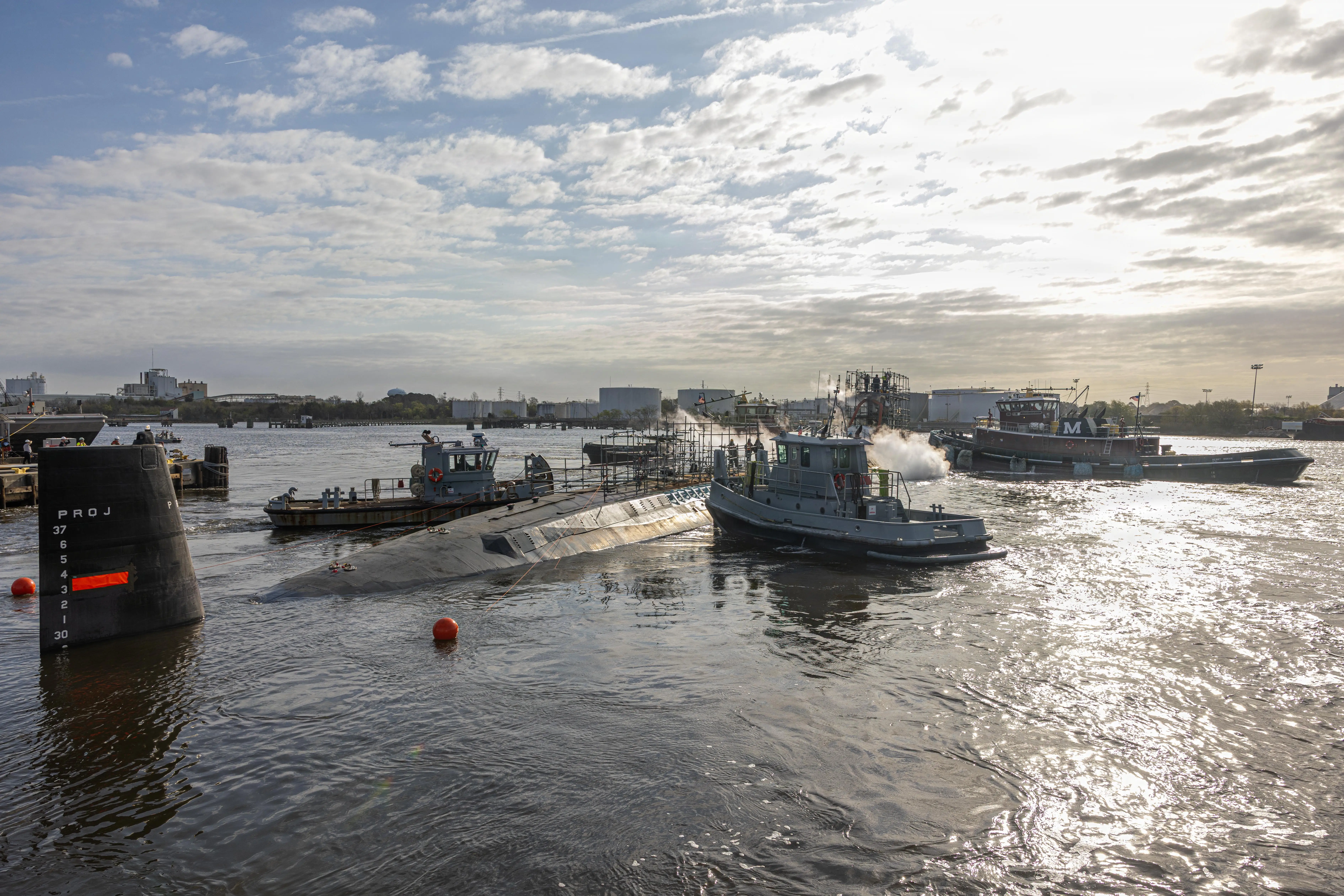 A submarine sits in dark blue water surrounded by several small boats. The sky is overcast in the background.