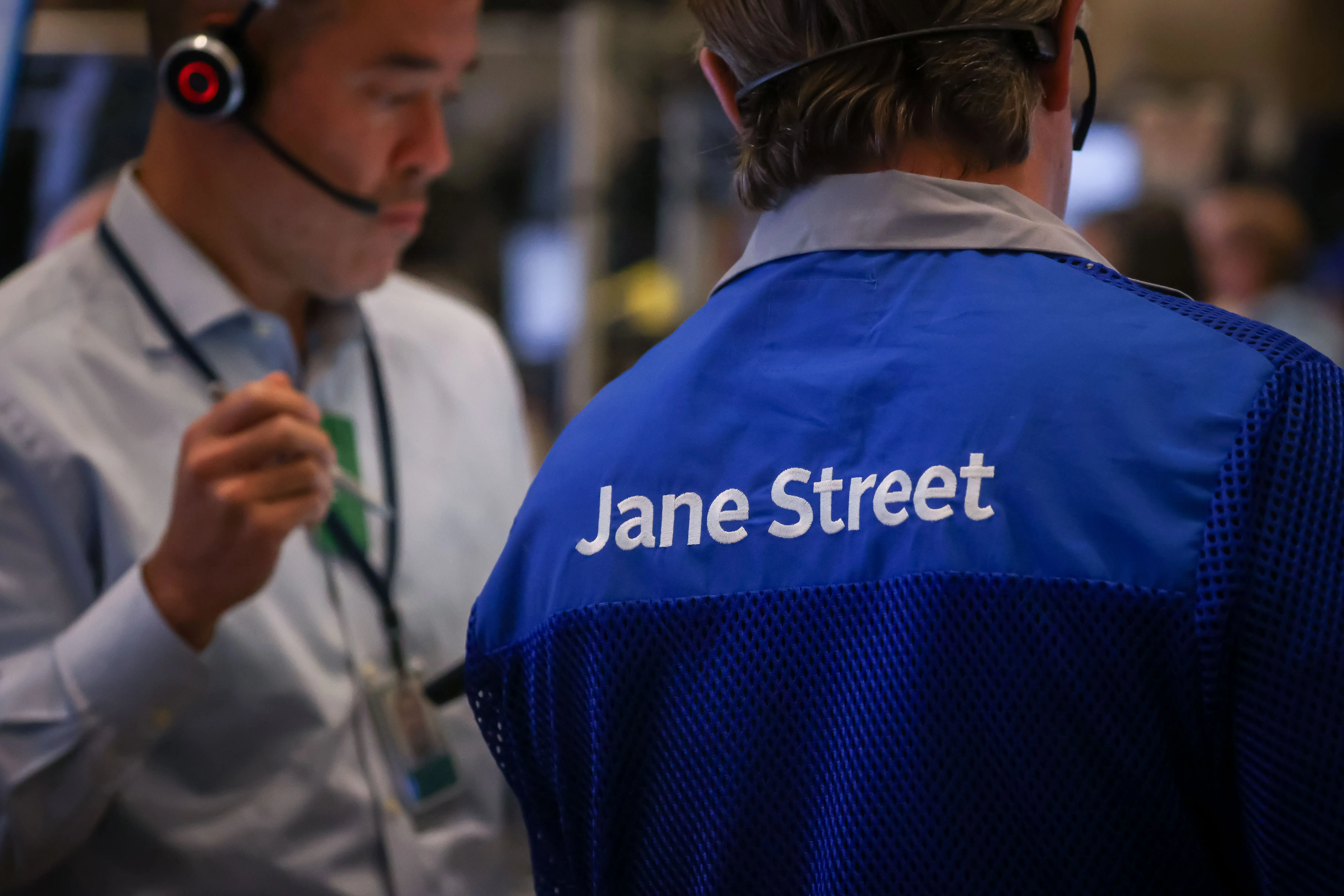 A trading floor worker in a blue vest with “Jane Street” on the back stands near headset-wearing staff.