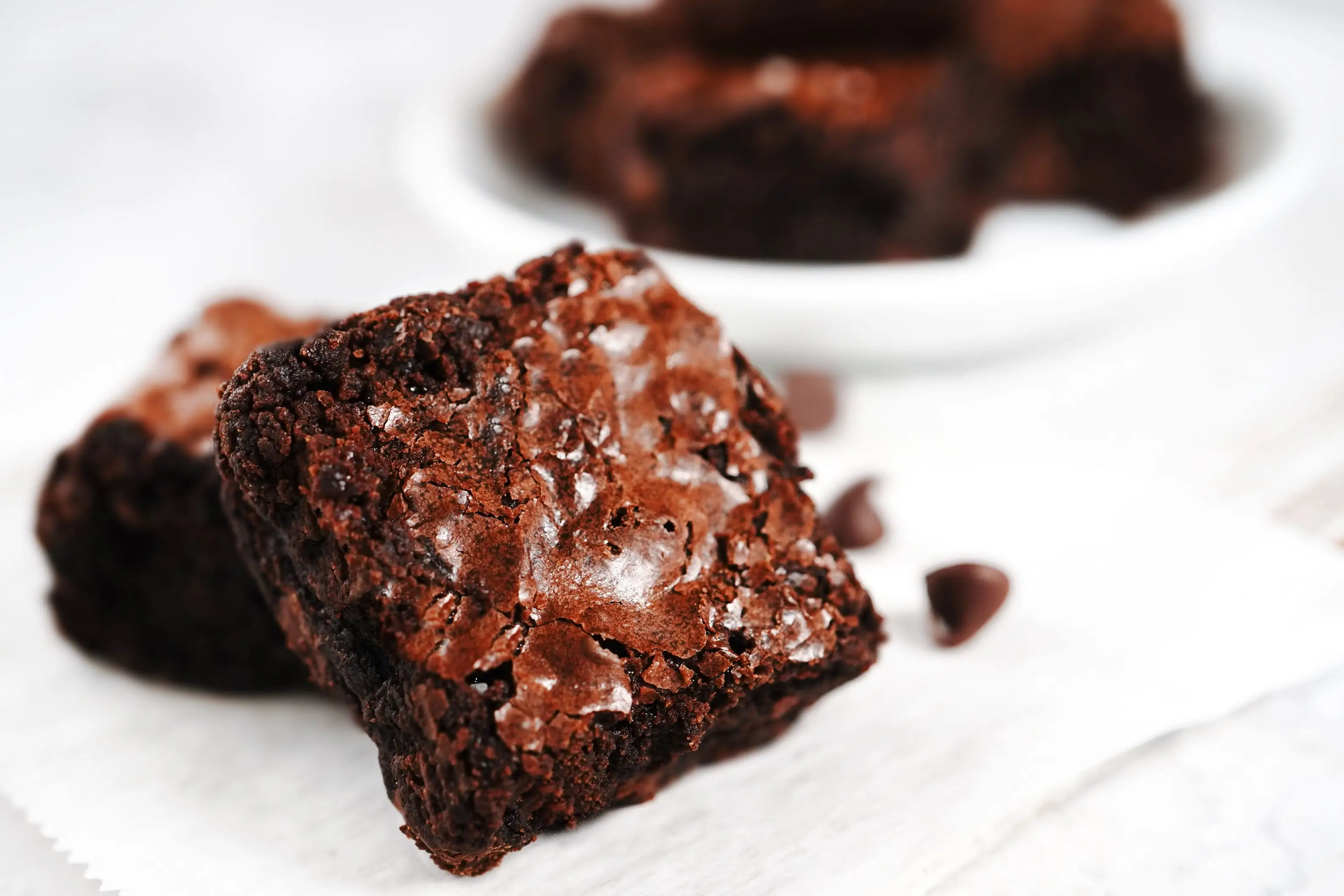 Chocolate brownies with glossy cracked tops sit on parchment paper with chocolate chips nearby.