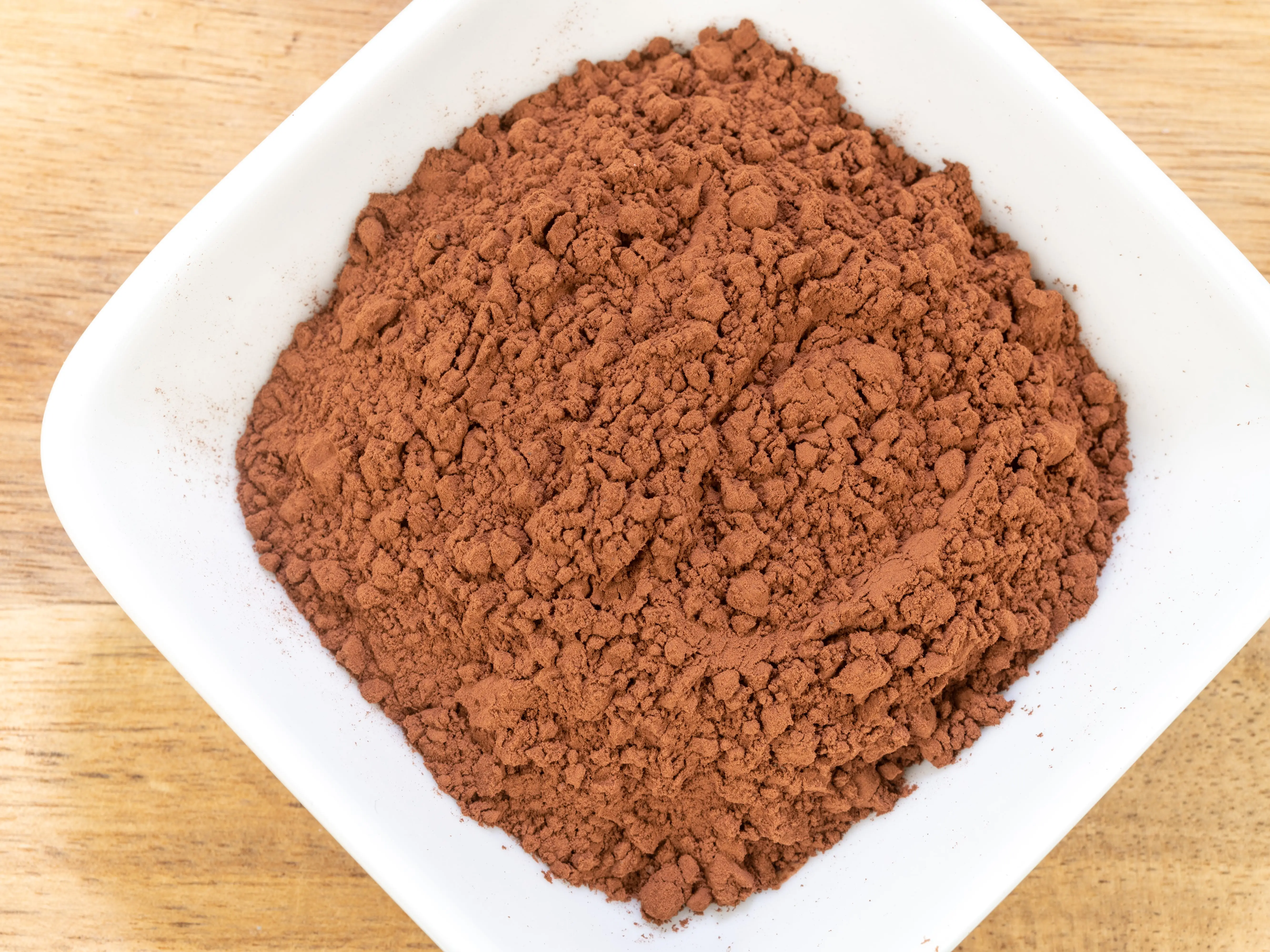 Brown cocoa powder fills a white square bowl on a light wooden surface.