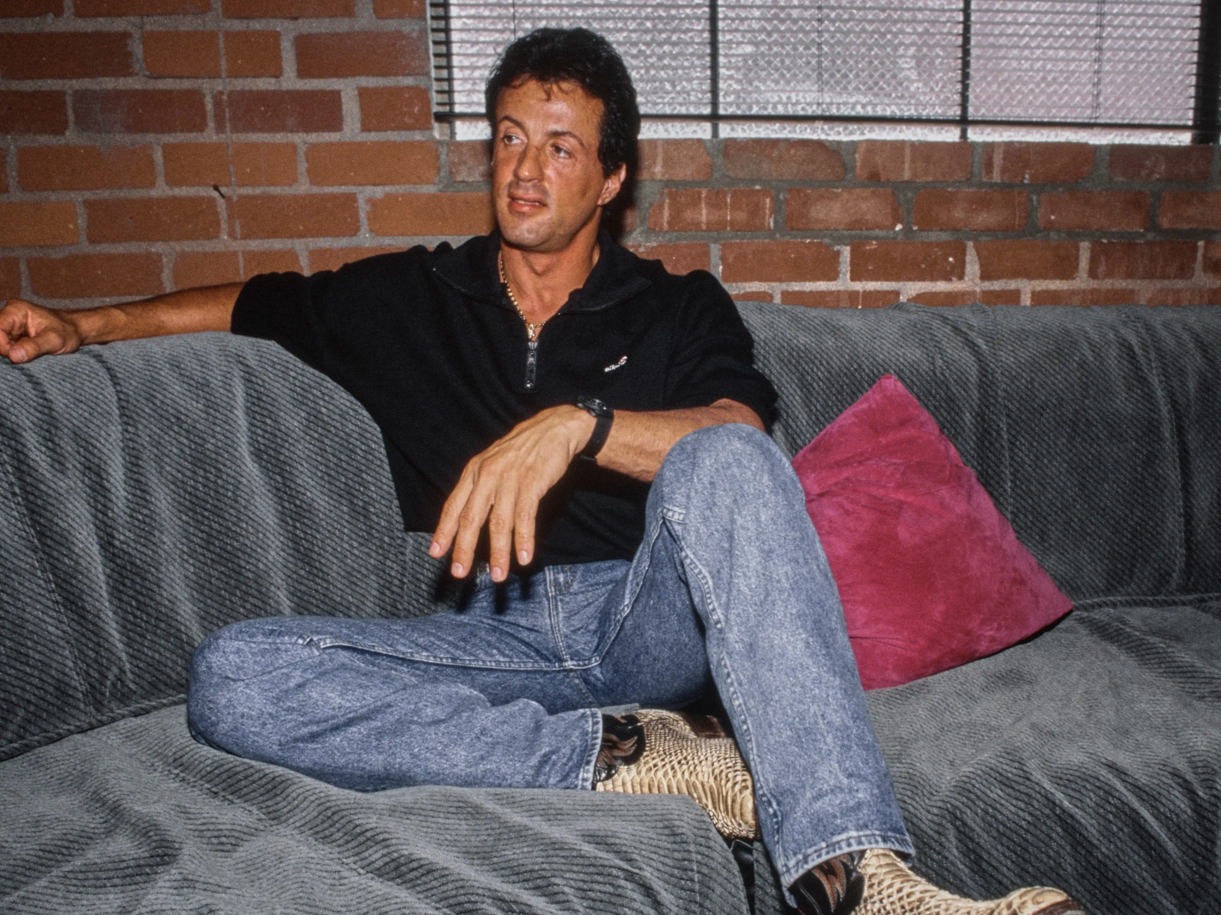 American actor Sylvester Stallone, wearing a black Ellesse polo shirt and stonewashed jeans with snakeskin boots, sitting beside a red cushion on a grey-and-black striped sofa, United States, circa 1982