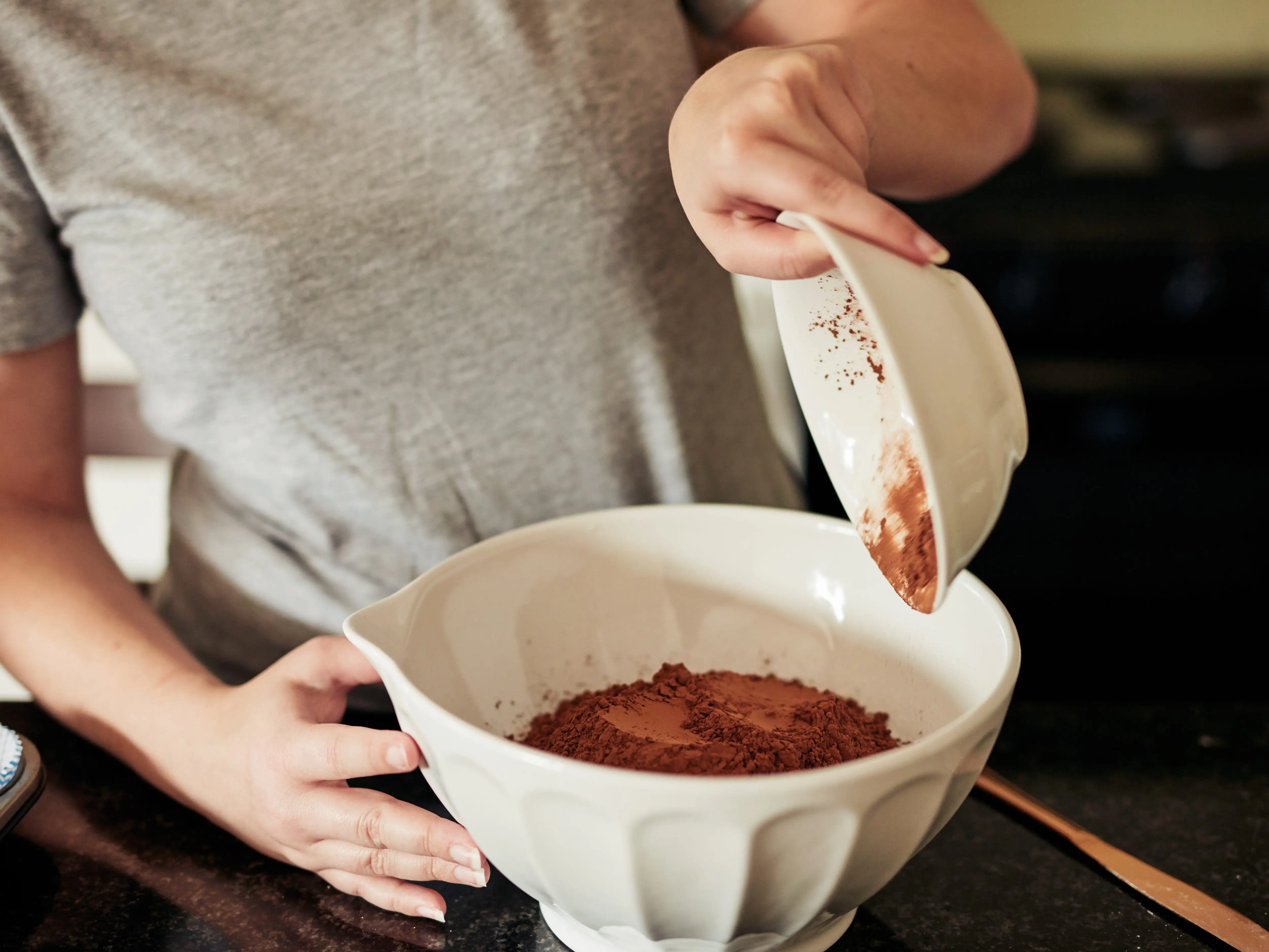 Hands pour brown powder from a small white bowl into a larger mixing bowl on a kitchen counter.