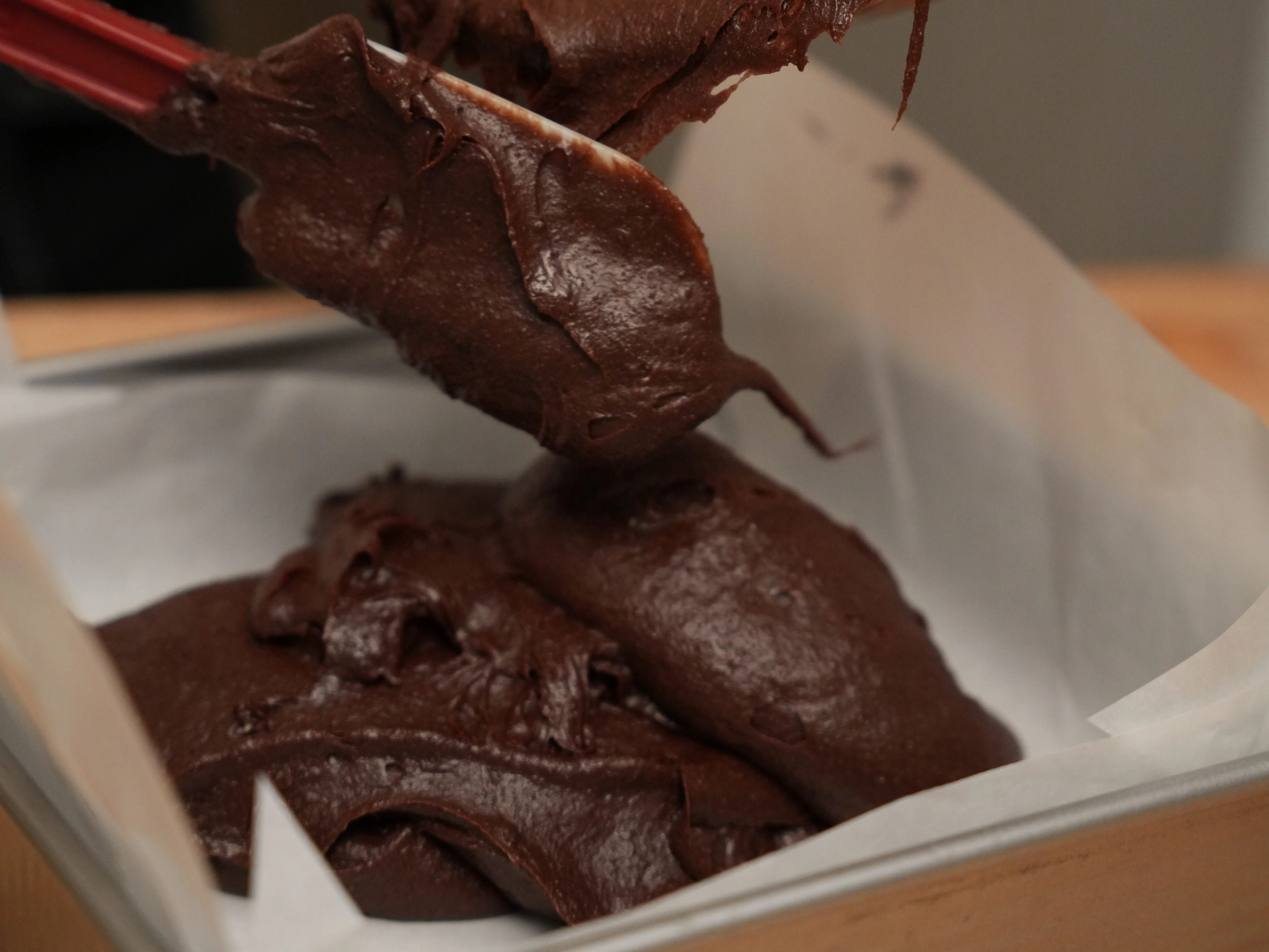 Thick chocolate batter is scraped from a mixing bowl into a parchment-lined square baking pan.