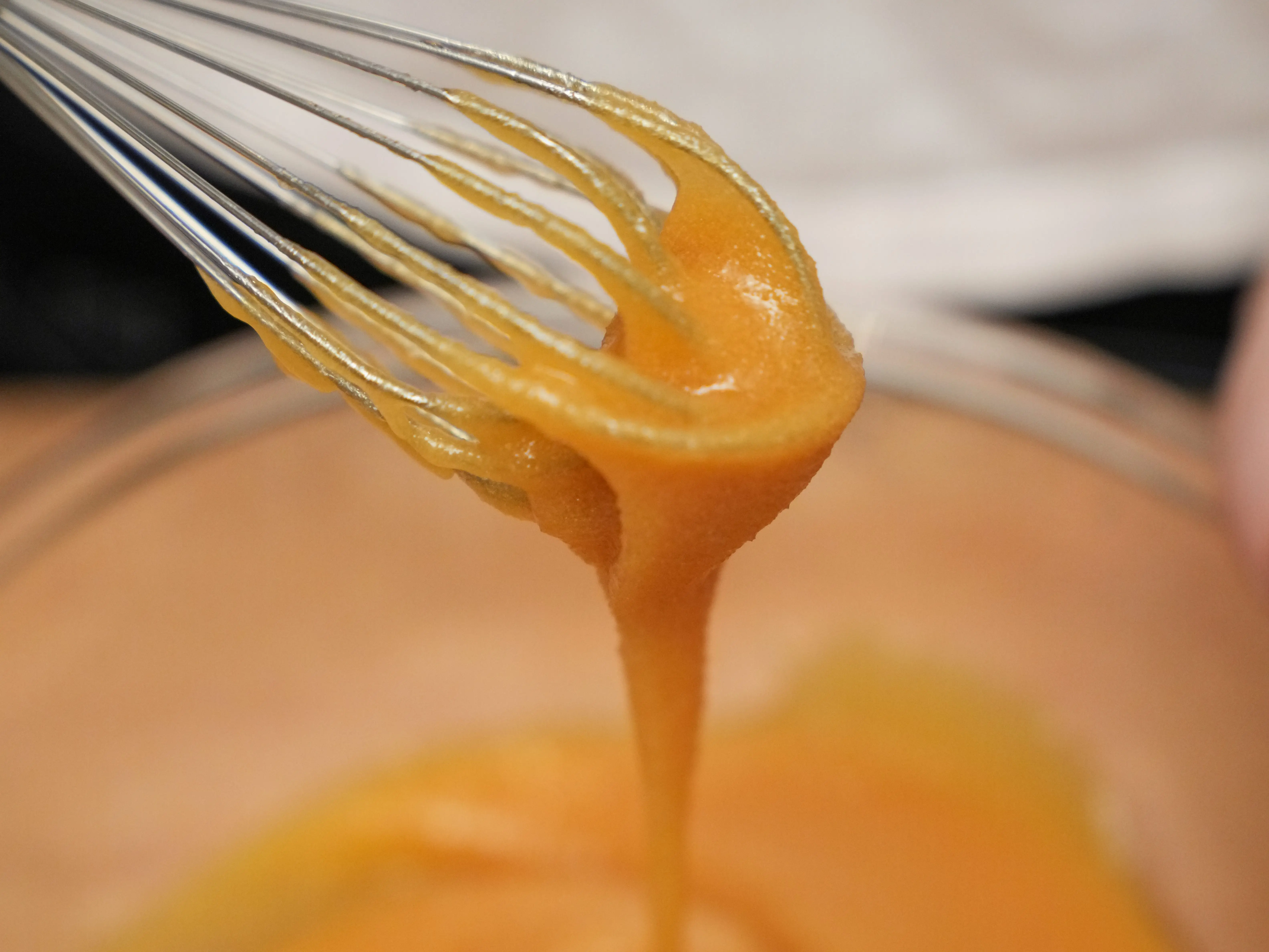 A metal whisk lifts a thick, pale-yellow-colored mixture as it drips back into a mixing bowl.