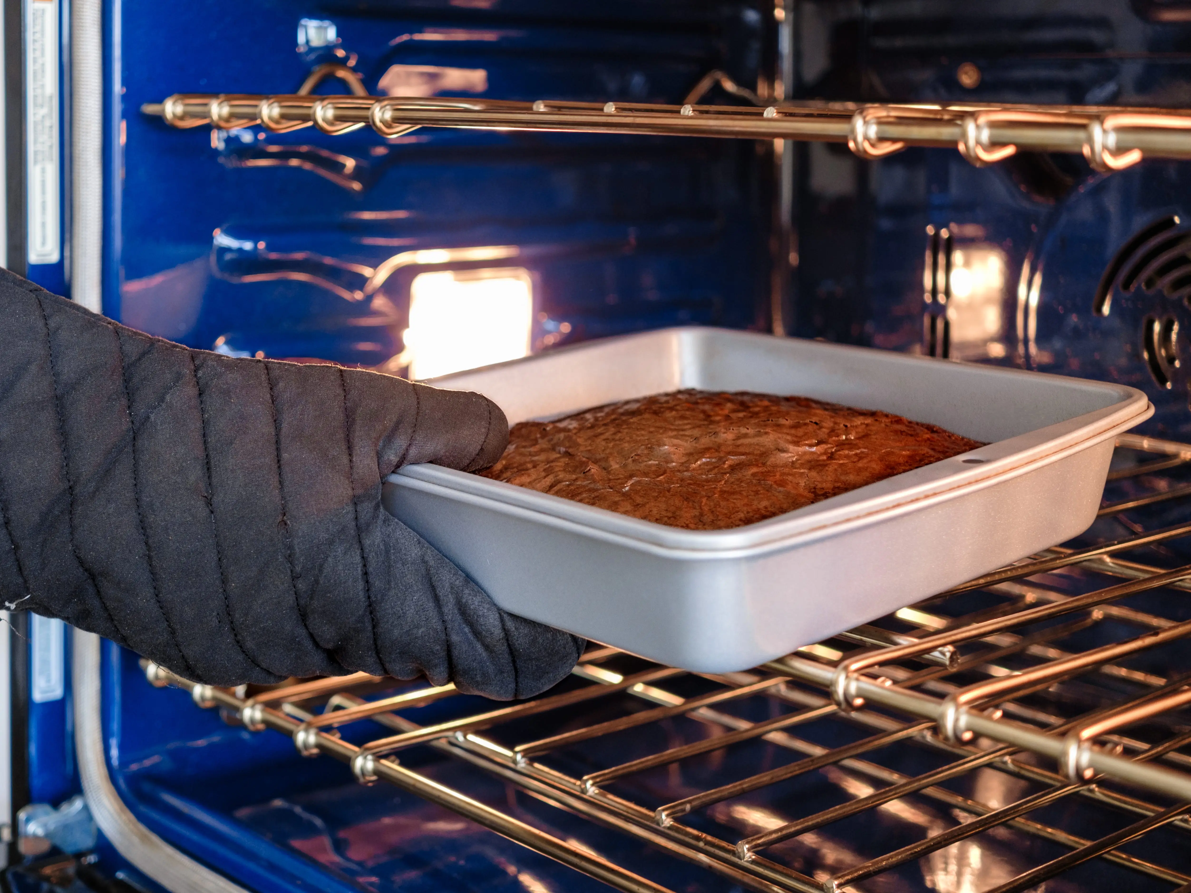 A gloved hand pulls a square pan of baked cake from a lit kitchen oven.