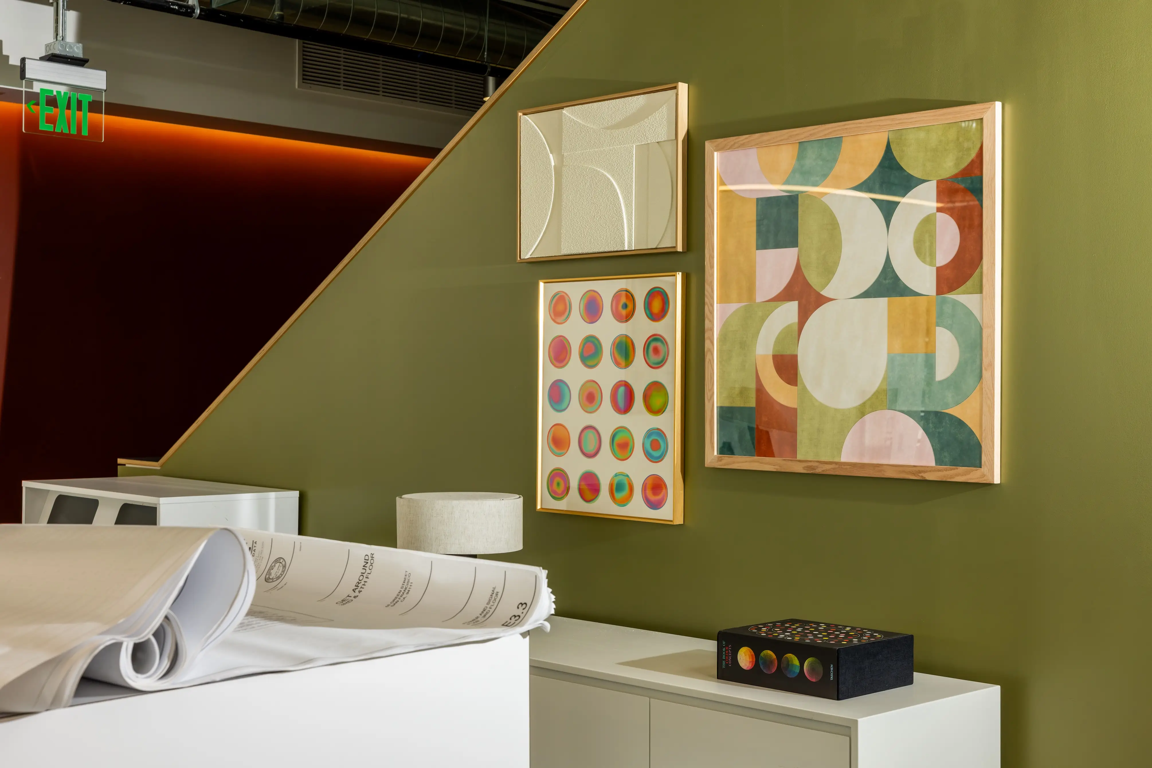 Framed abstract artworks hang on an olive green wall above cabinets and rolled architectural plans.