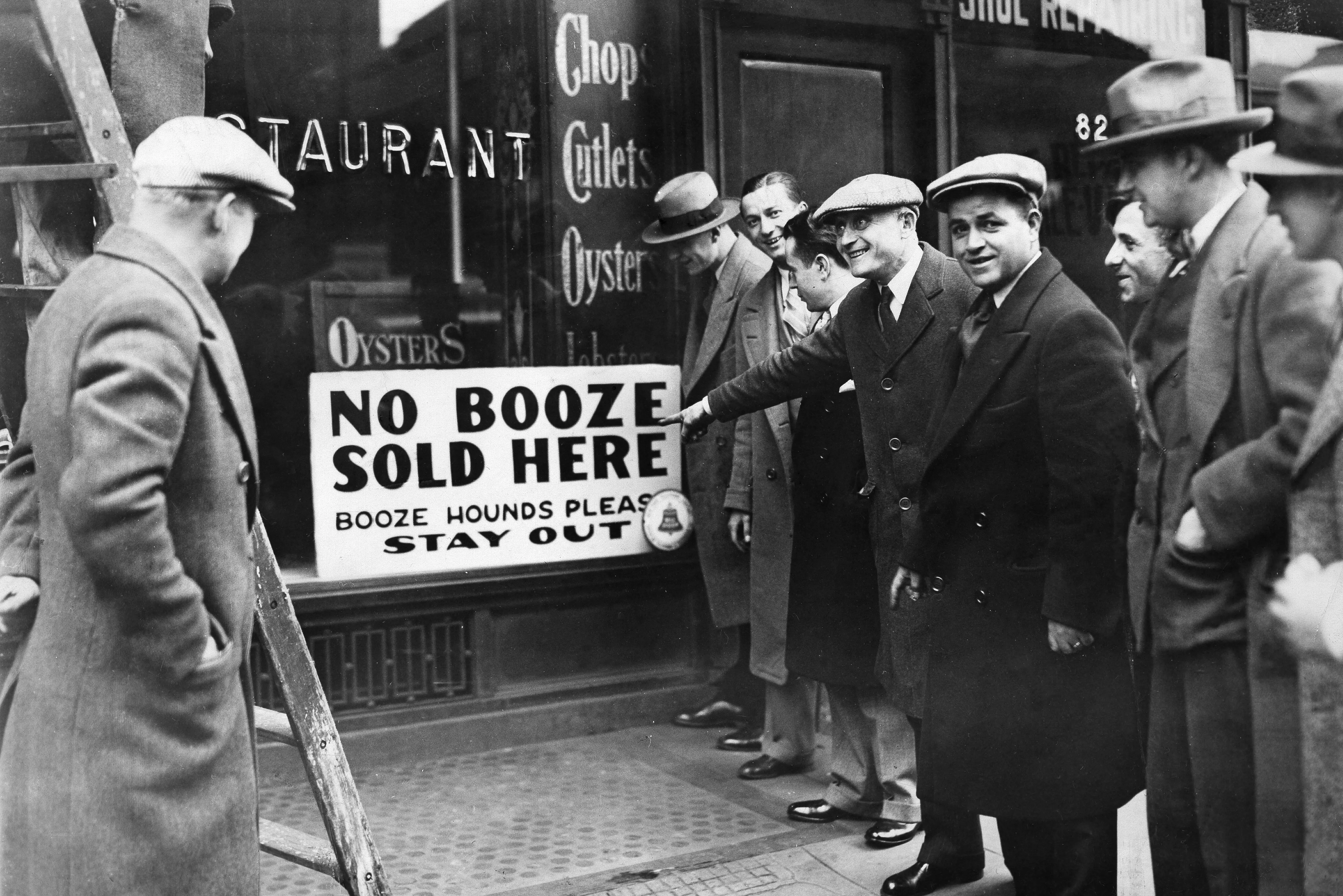 People in hats gather outside a restaurant beside a sign reading “NO BOOZE SOLD HERE.”