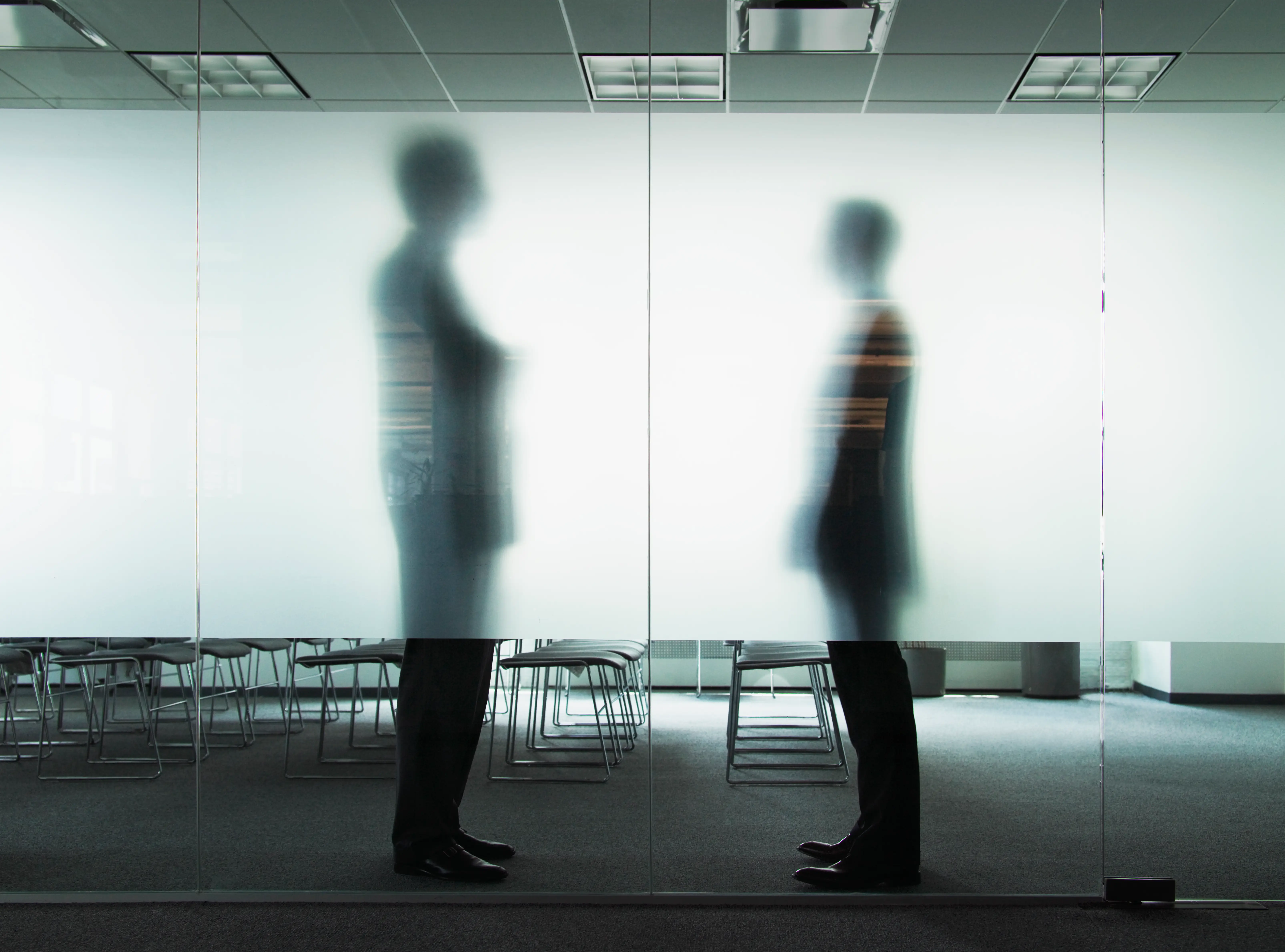 Two blurred silhouettes stand facing each other behind frosted glass in a modern office meeting room.