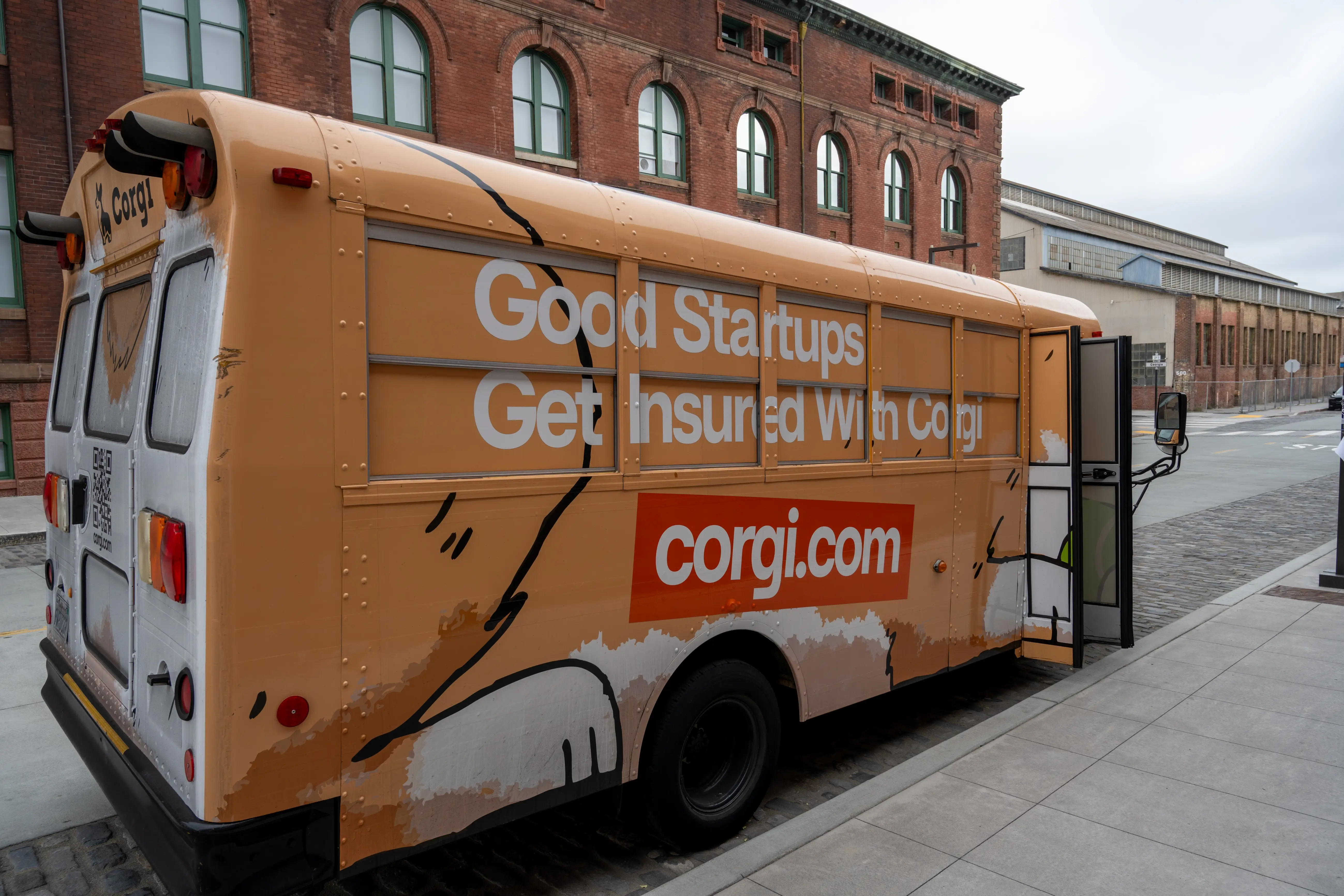 Orange corgi.com branded bus parked on a city street beside brick industrial buildings.