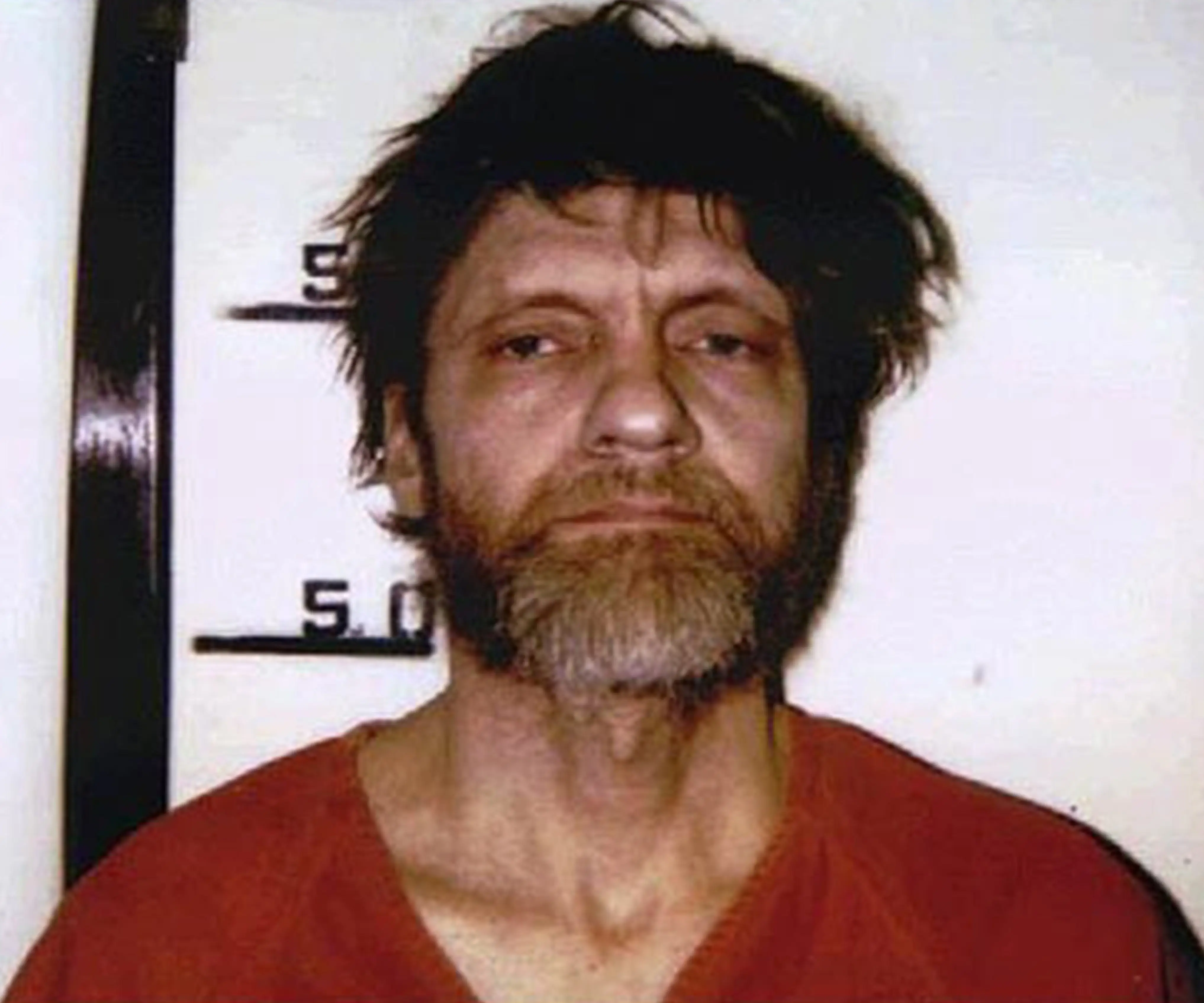 Ted Kaczynski faces the camera in a red shirt against a height chart backdrop.