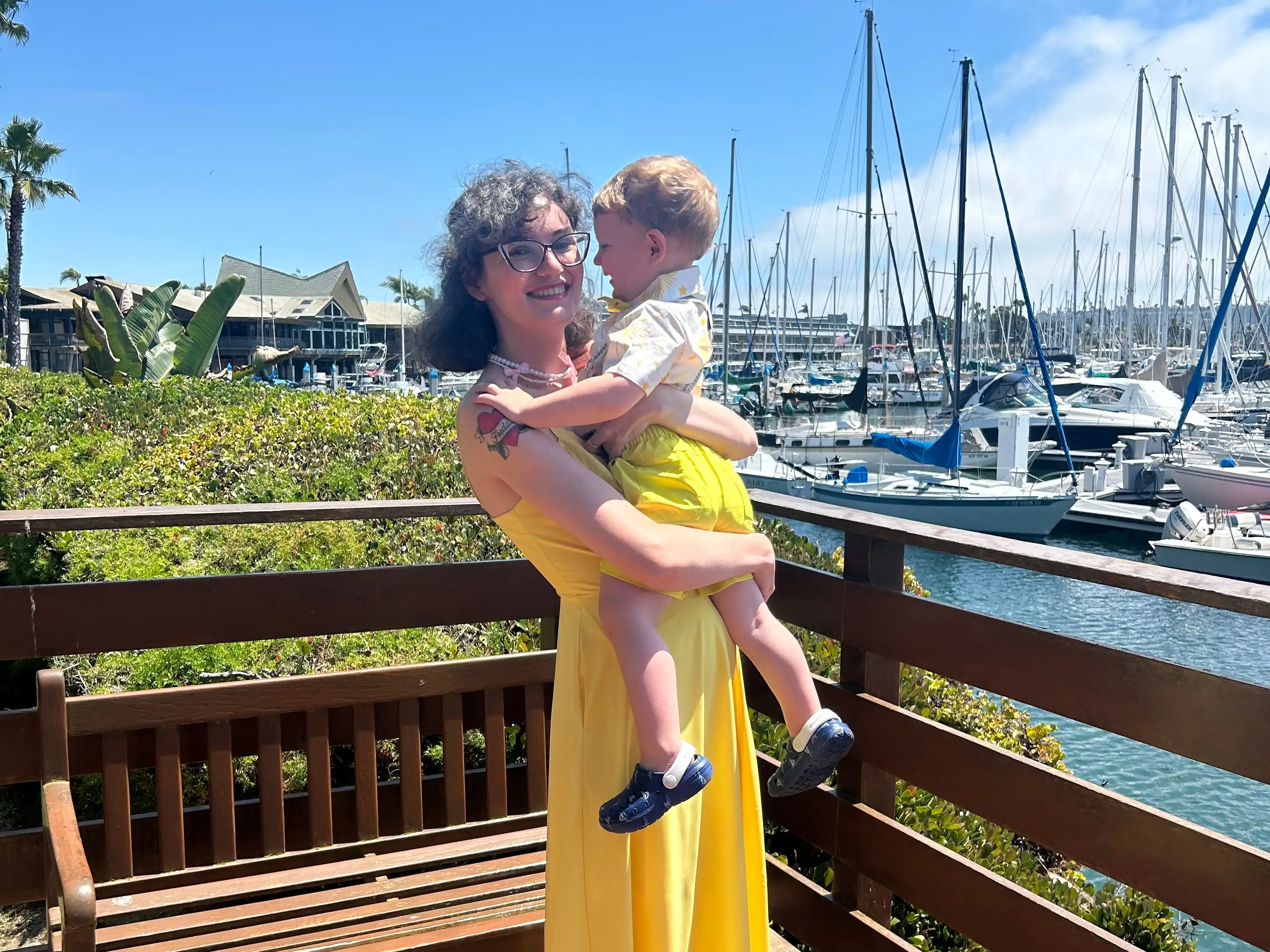 Person in a yellow dress holds a small child on a wooden deck overlooking a marina with sailboats.
