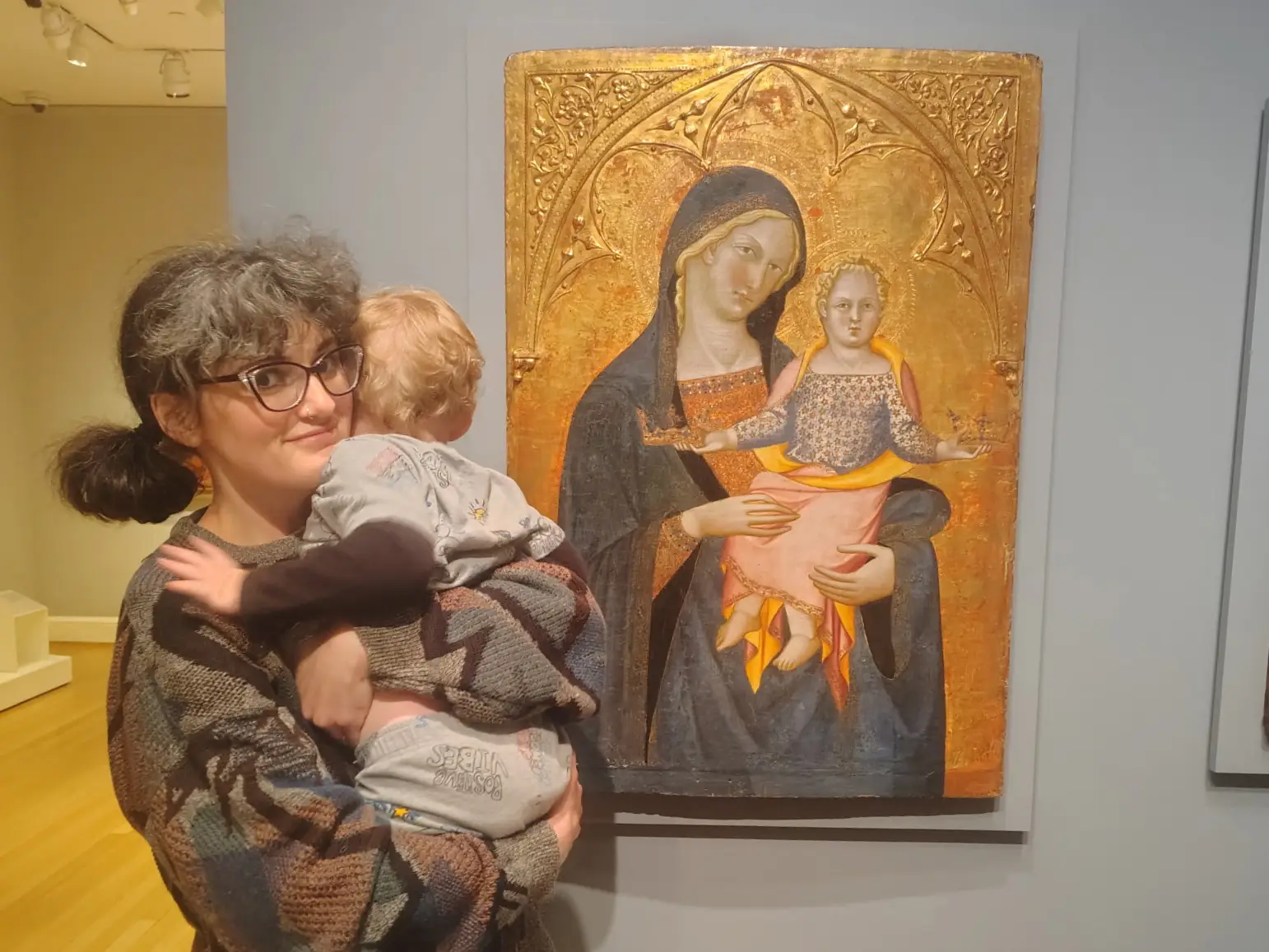Person wearing glasses holds a small child beside a gilded Madonna and Child painting in a museum gallery.