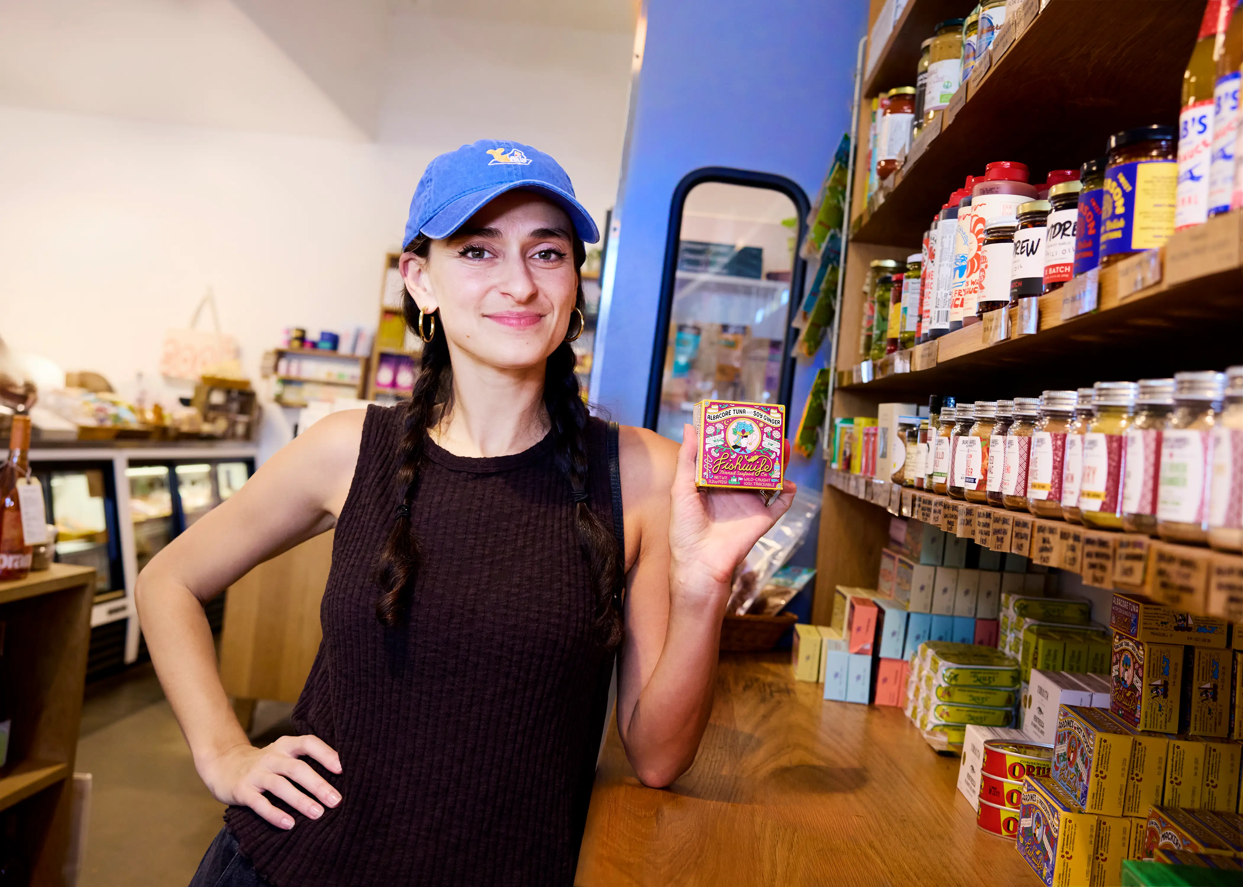 Person in a blue cap holds a colorful canned food package beside stocked specialty shop shelves.