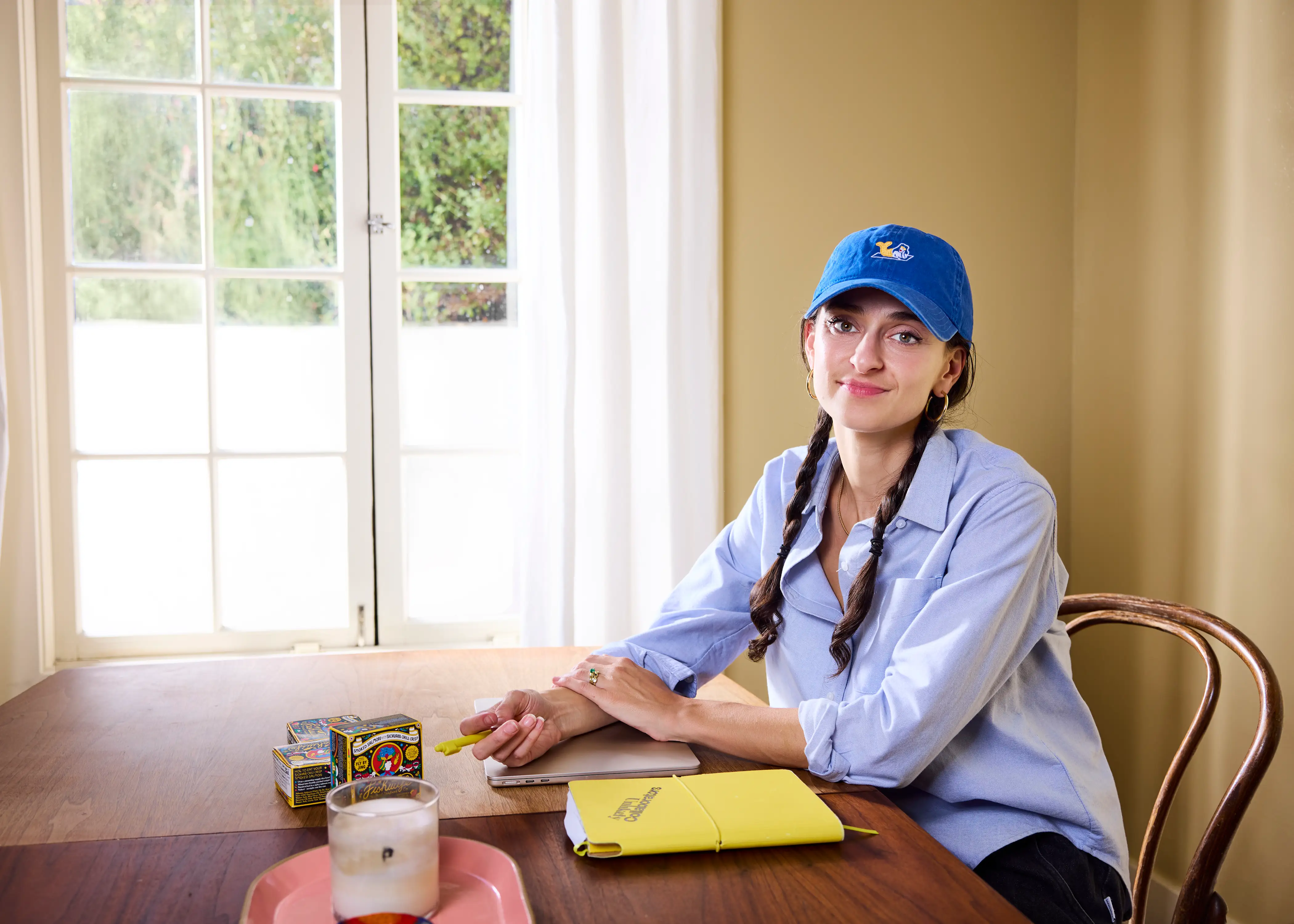Person in a blue cap sits at a wooden table with stationery, boxes, and a drink near tall windows.