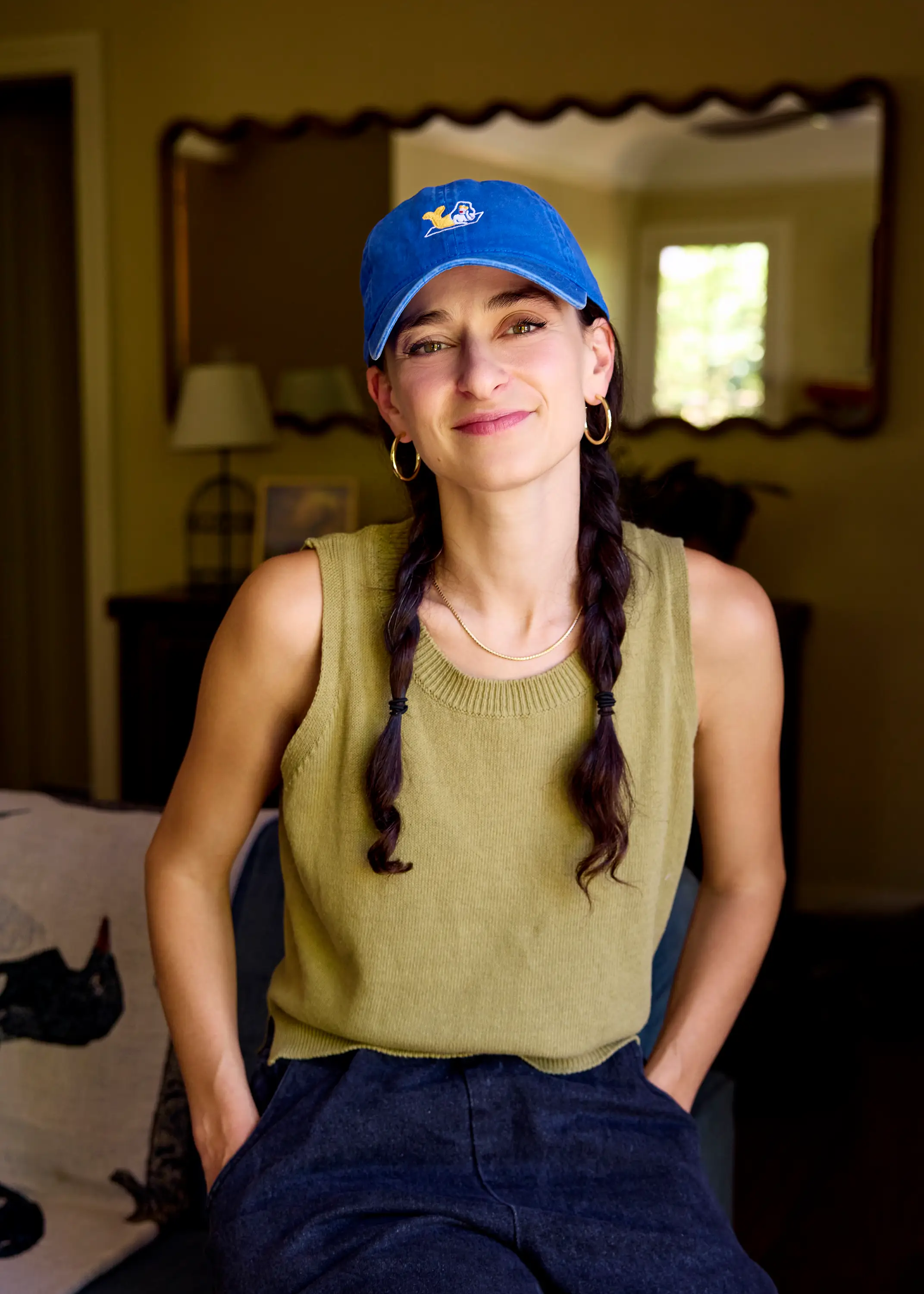 Person wearing a blue baseball cap and green sleeveless knit top sits indoors in front of a mirror.
