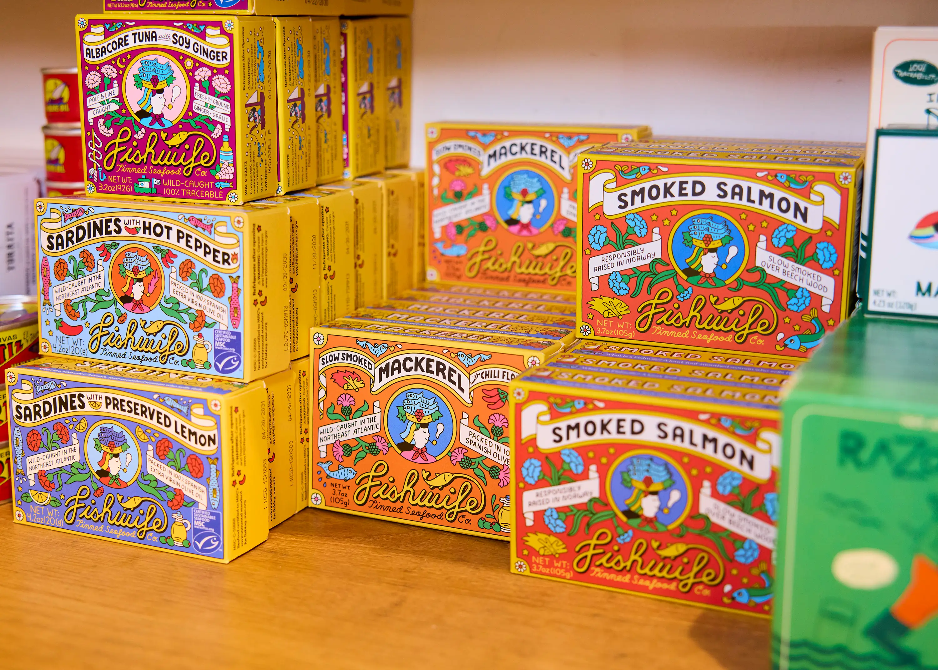Colorful Fishwife tinned seafood boxes are stacked on a wooden store shelf among other packaged products.