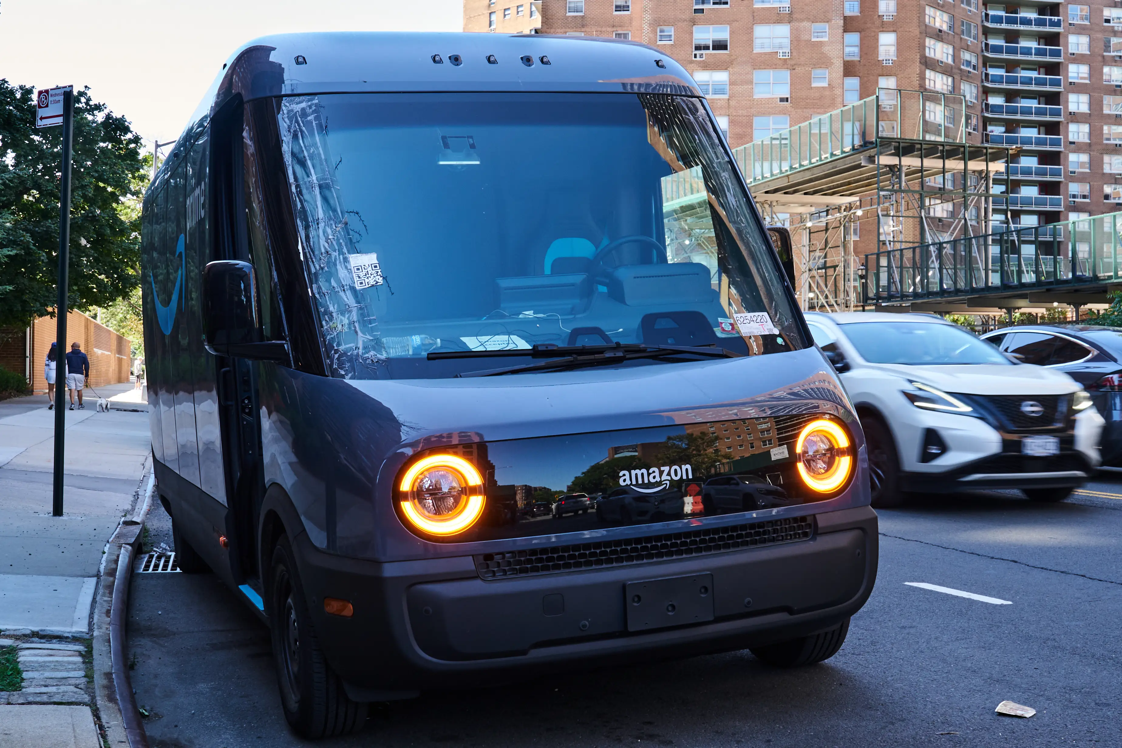 Amazon-branded delivery van with illuminated headlights parked at a curb on a city street.