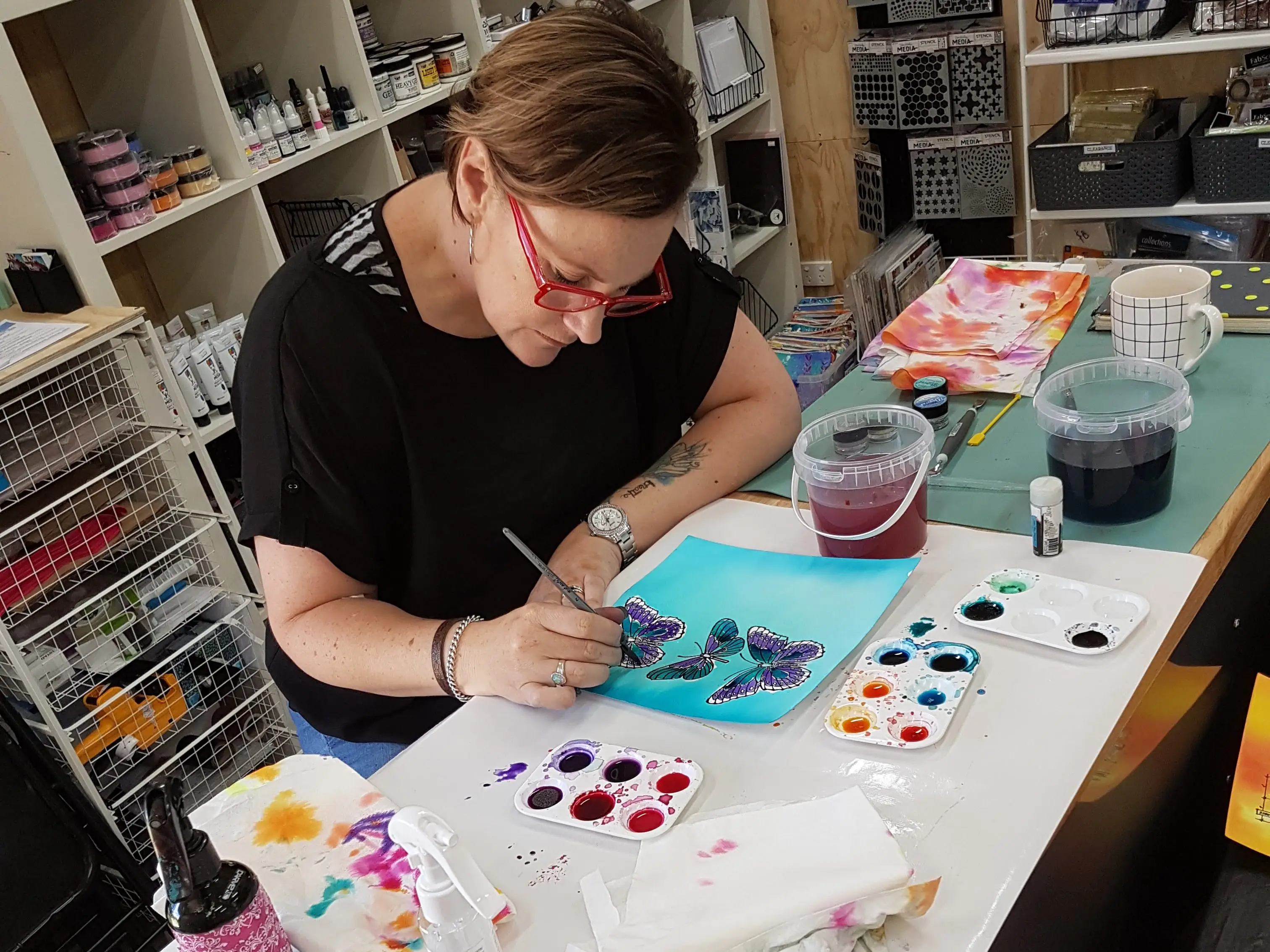 Person wearing red glasses paints a butterfly design at a craft table surrounded by inks and art supplies.