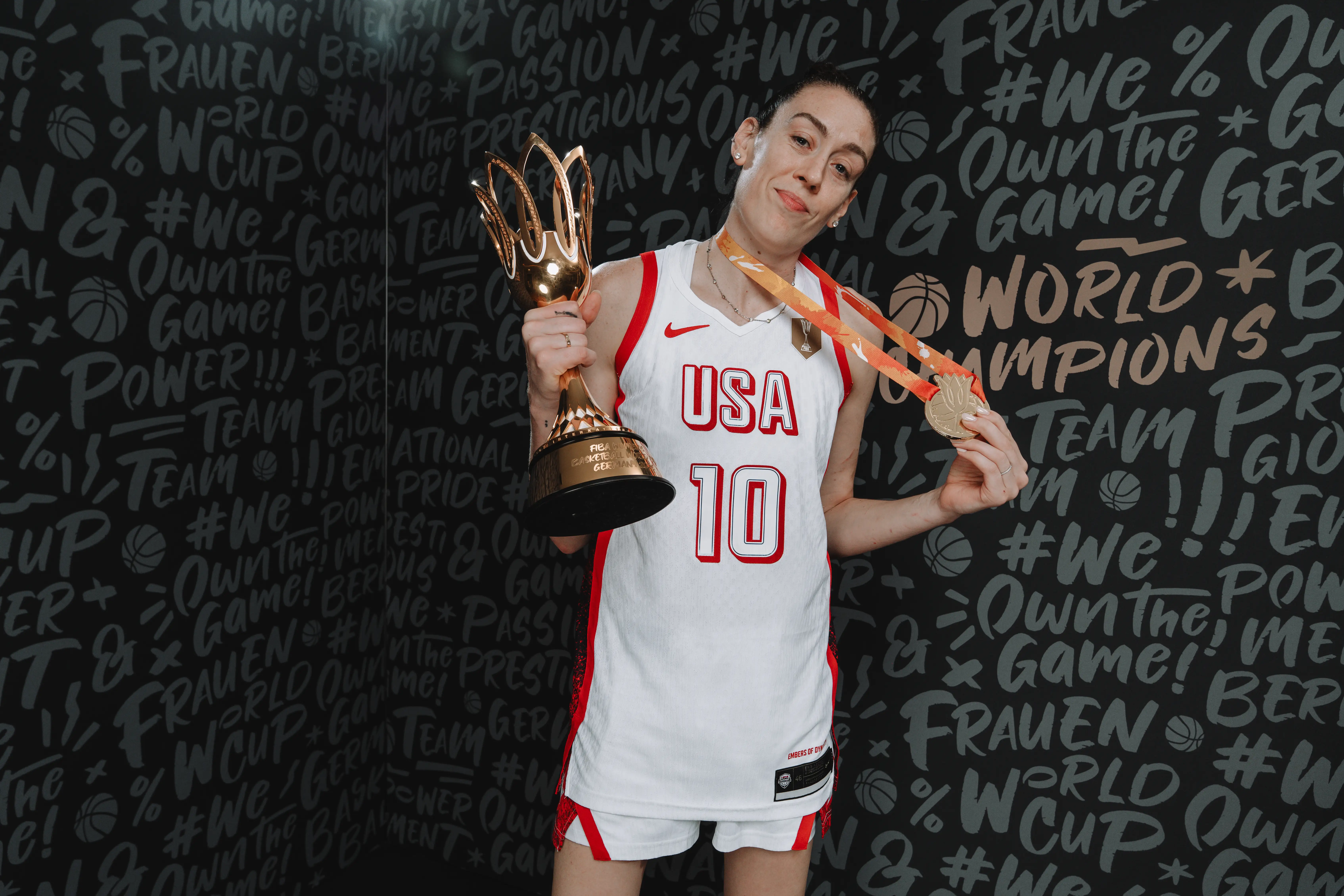 Breanna Stewart poses in a USA basketball jersey holding a trophy and medal before a championship-themed backdrop.